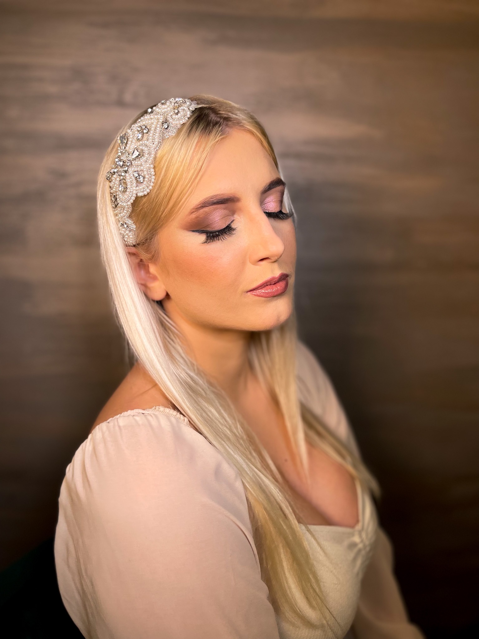 Portret kobiety z blond włosami i ozdobną opaską, z zamkniętymi oczami i subtelnym, różowym cieniem na powiekach oraz delikatnym błyszczykiem na ustach.