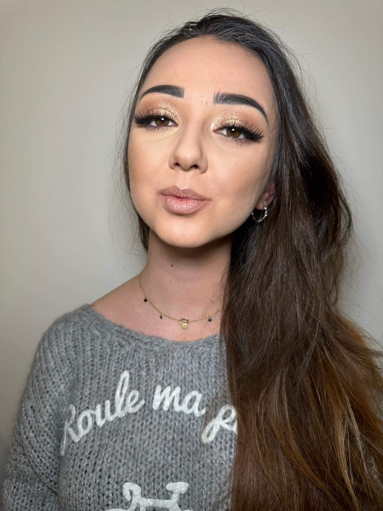 Portret kobiety z błyszczącym, złotym cieniem na powiekach i długimi, ciemnymi włosami, ubrana w szary sweter z napisem.