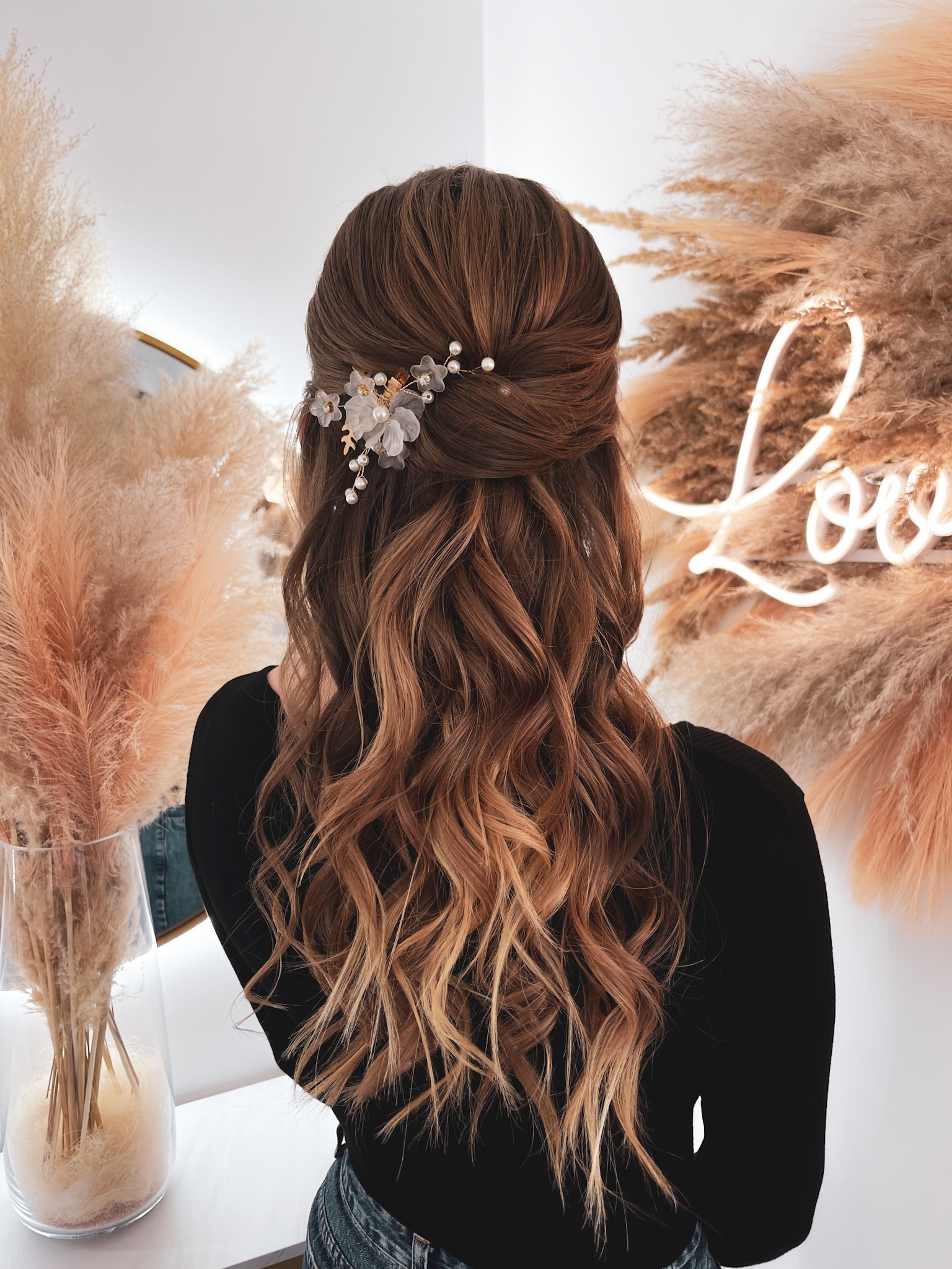 Uczesanie z podpiętymi włosami, ozdobione delikatną spinką z kwiatami i perłami, na tle dekoracji z pampasów i neonowego napisu.
