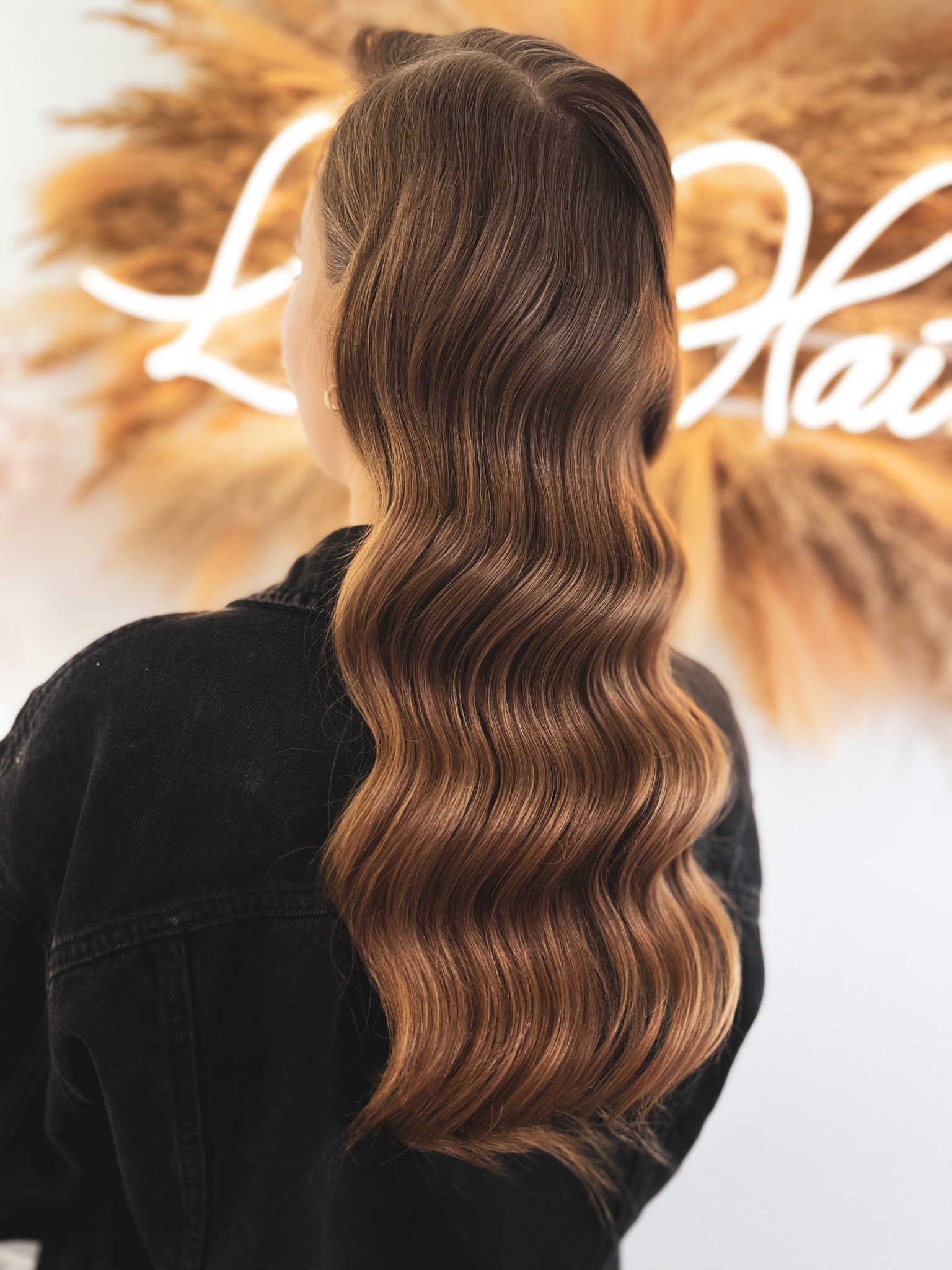 Brązowe, falowane włosy opadające na plecy kobiety ubranej w czarną kurtkę dżinsową. W tle dekoracja ze sztucznych traw i neonowy napis.