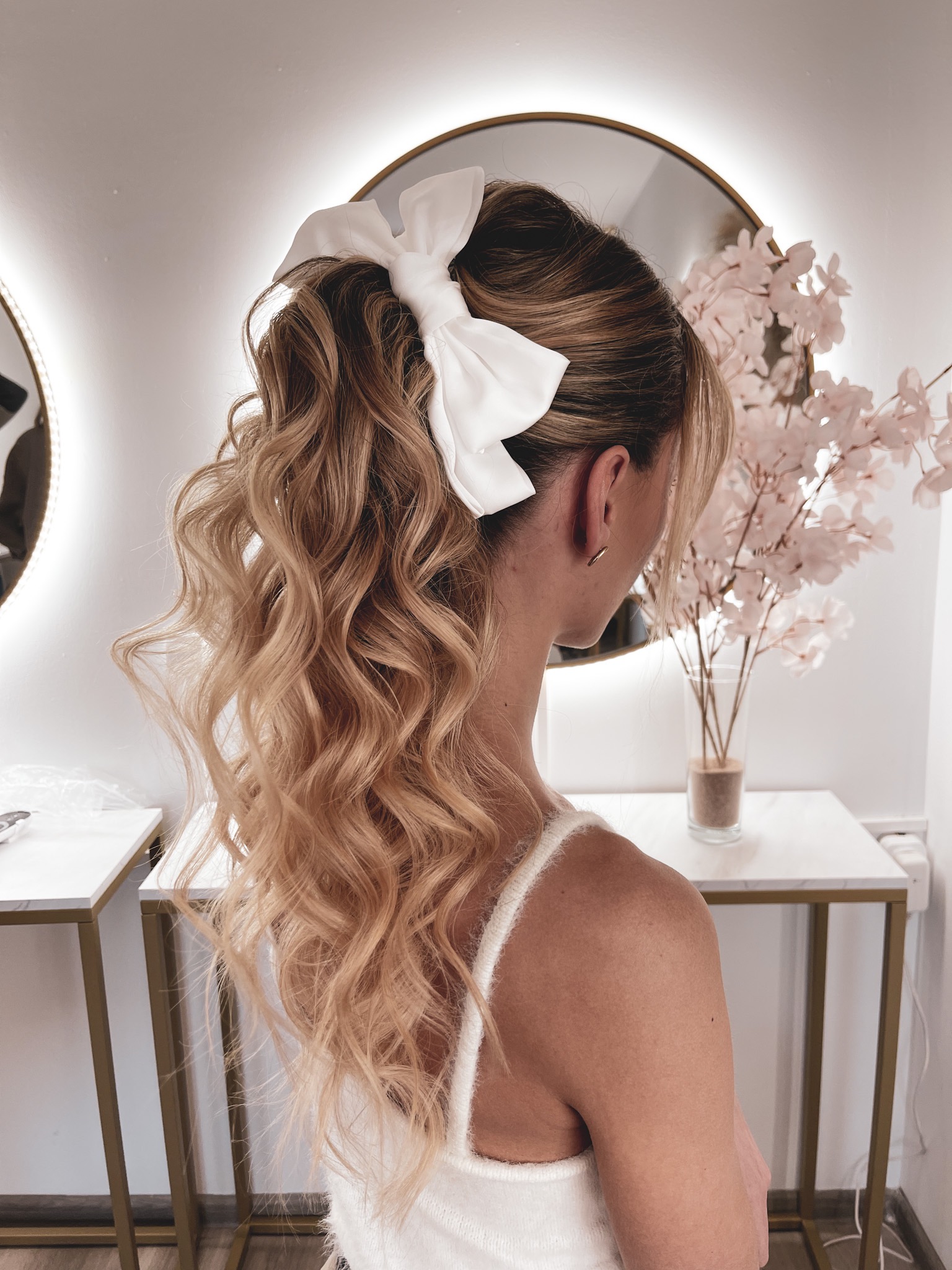 Upięcie włosów w Szczecinie: blond loki zebrane w wysoki kucyk, ozdobione dużą białą kokardą, widok z tyłu, delikatny makijaż, minimalistyczne kolczyki.