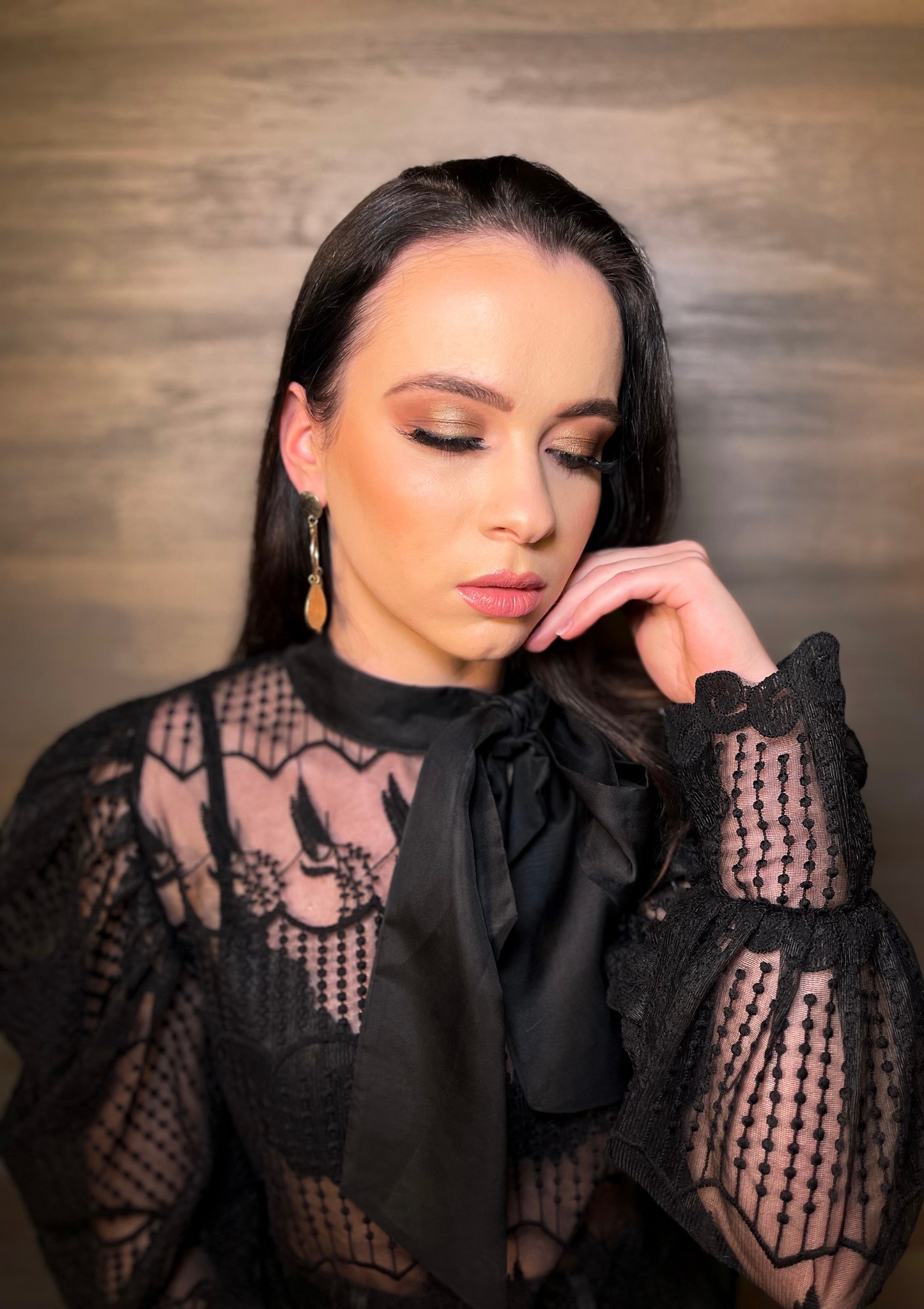 Portret kobiety z zamkniętymi oczami, prezentującej złoty makijaż oczu i subtelny makijaż ust, ubrana w czarną, koronkową bluzkę z kokardą, z wiszącymi kolczykami na tle drewnianej ściany.