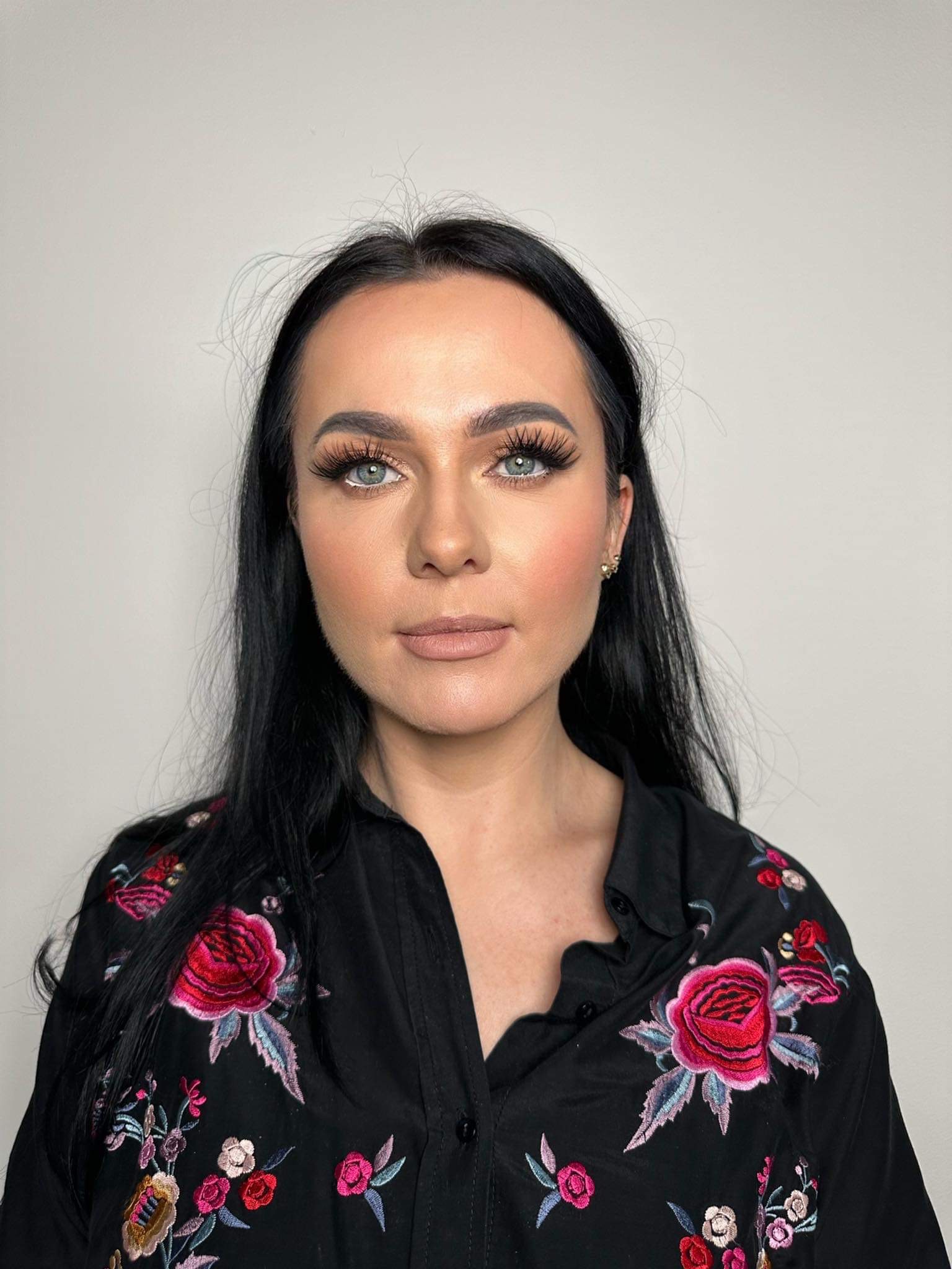 Portret kobiety z pełnym, naturalnym makijażem, podkreślonymi rzęsami i delikatnym konturowaniem, ubrana w czarną koszulę z haftowanymi różami.