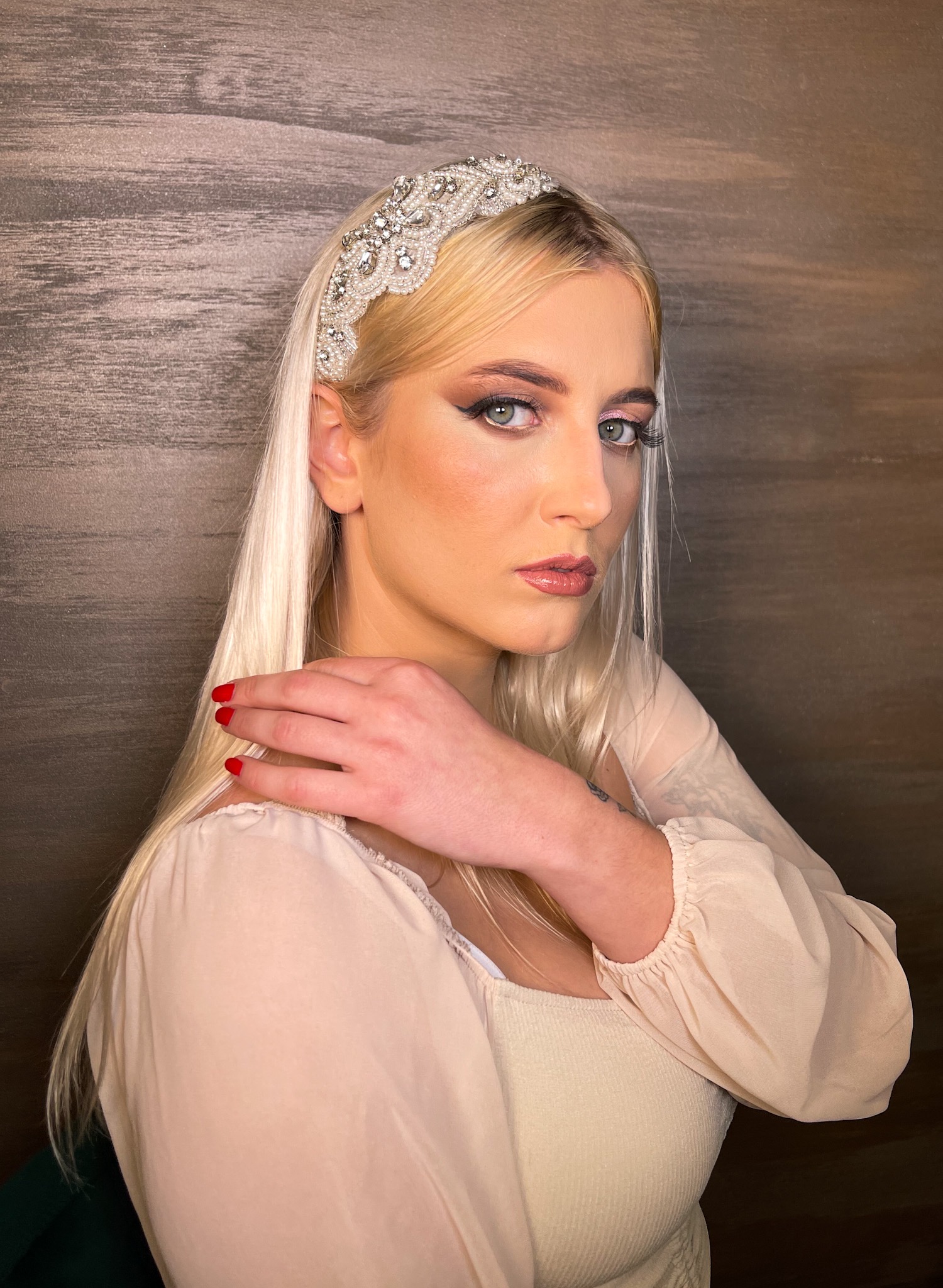 Portret kobiety z platynowymi włosami, w ozdobnej opasce wysadzanej kamieniami, z wyraźnym makijażem oczu i ust, pozującej na tle ściany o fakturze drewna.