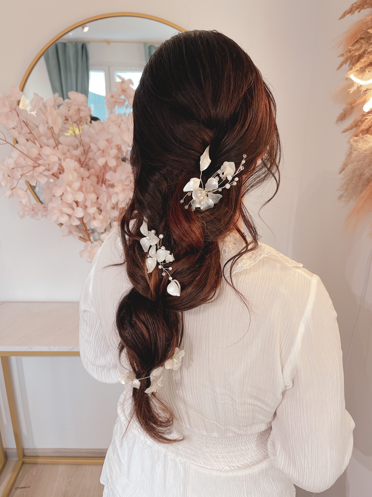 Upięcie włosów w warkocz z dekoracyjnymi, białymi spinkami w kształcie kwiatów i liści, widok z tyłu, na tle lustra i dekoracji kwiatowych.