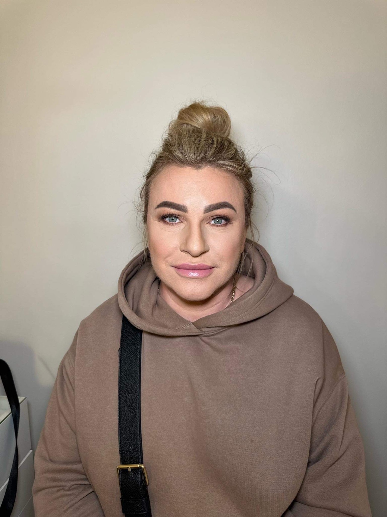 Portret kobiety z upiętymi blond włosami, delikatnym makijażem i w brązowej bluzie z kapturem, na neutralnym tle.