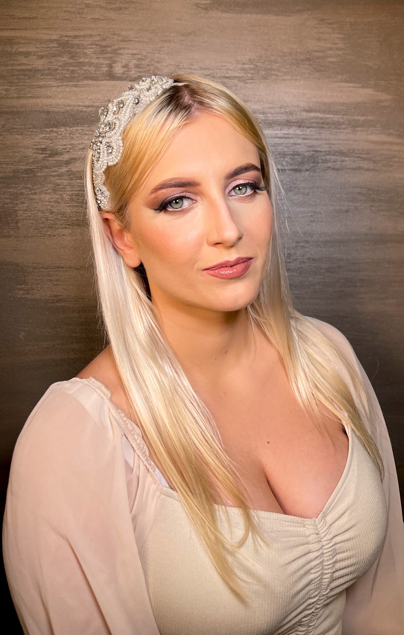 Portret blondynki z delikatnym, różowym makijażem oczu i ust, zdobioną opaską z pereł i kryształów na włosach na tle drewnianej ściany.