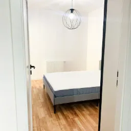 Widok na sypialnię z łóżkiem na drewnianej podłodze przez otwarte drzwi, wisząca lampa w stylu industrialnym i białe ściany.