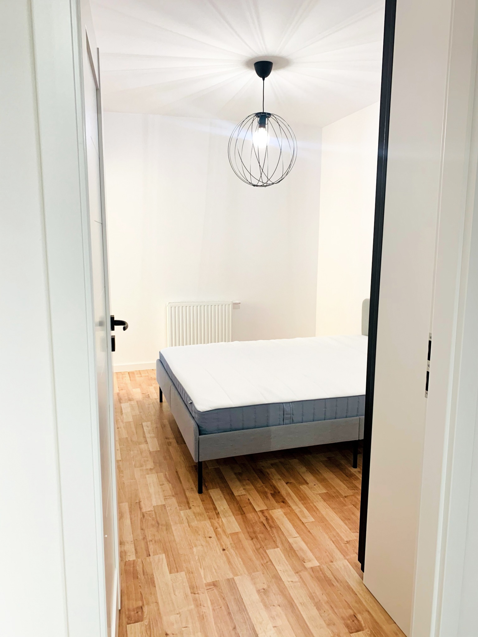 Widok na sypialnię z łóżkiem na drewnianej podłodze przez otwarte drzwi, wisząca lampa w stylu industrialnym i białe ściany.