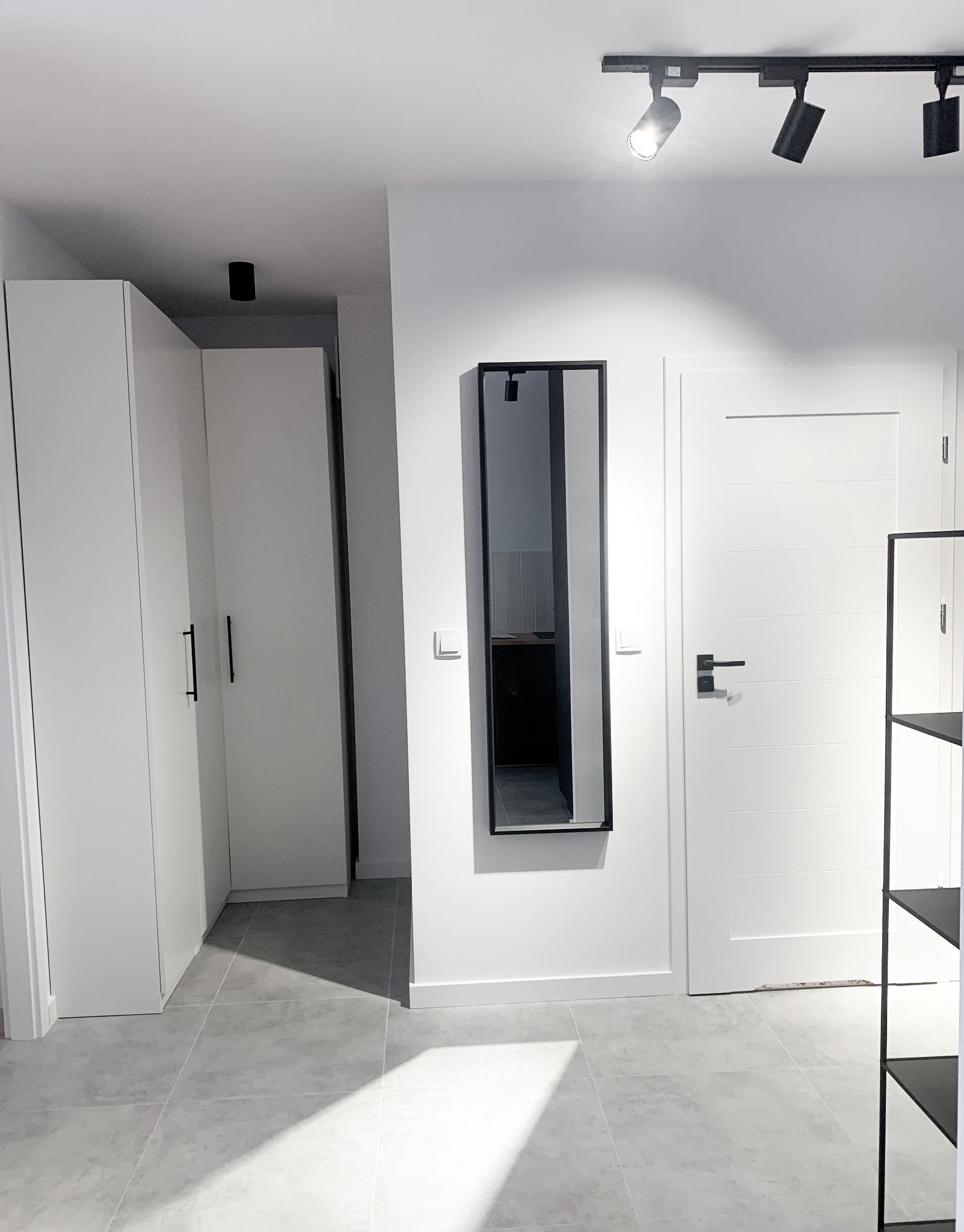 Nowoczesne wnętrze przedpokoju z szarą płytką podłogową, białymi szafami z czarnymi uchwytami, lustrem w czarnej ramie i oświetleniem punktowym.