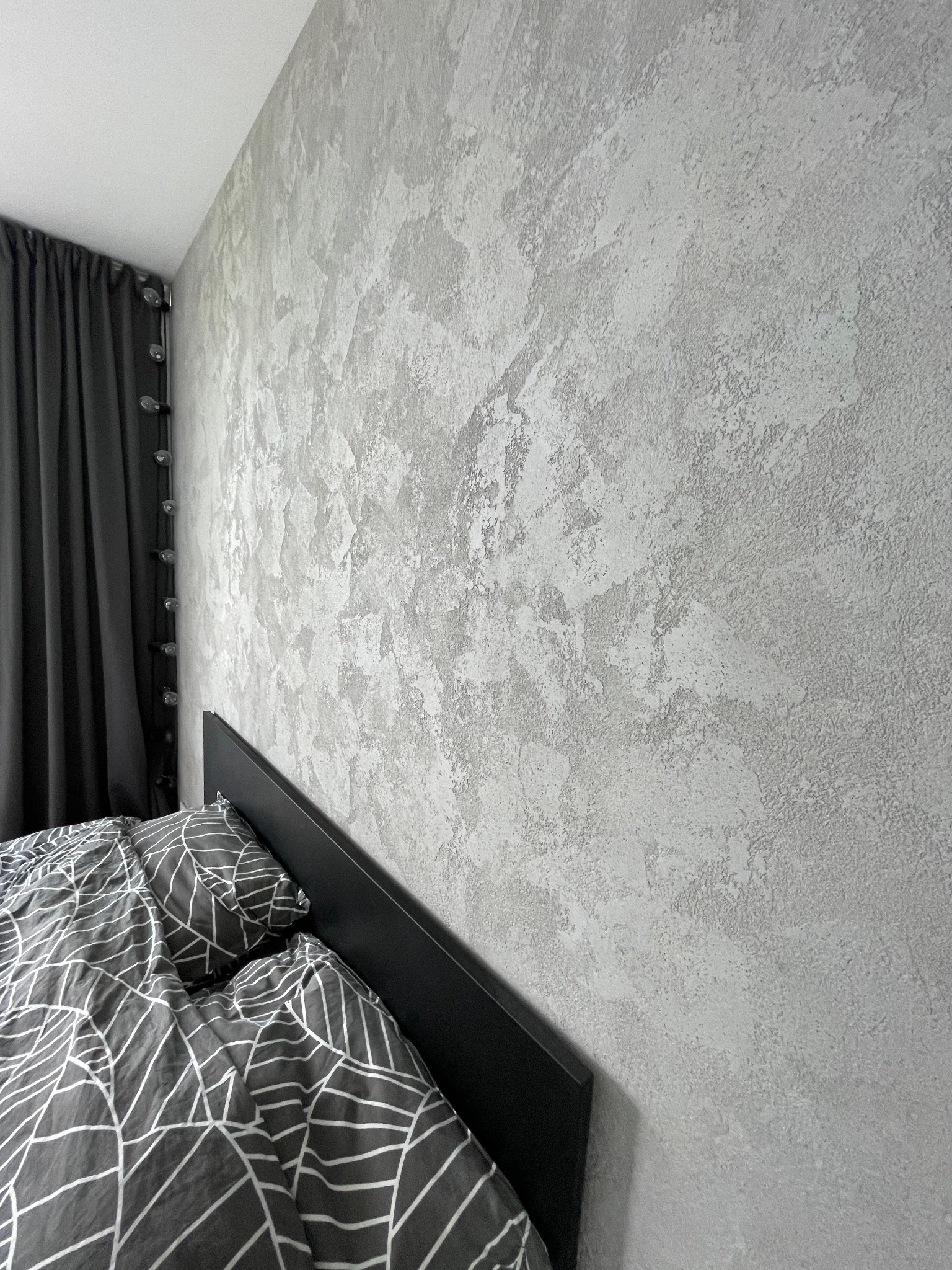 Szara ściana z efektem dekoracyjnym, widoczna część łóżka z pościelą w geometryczne wzory oraz fragment zasłony.