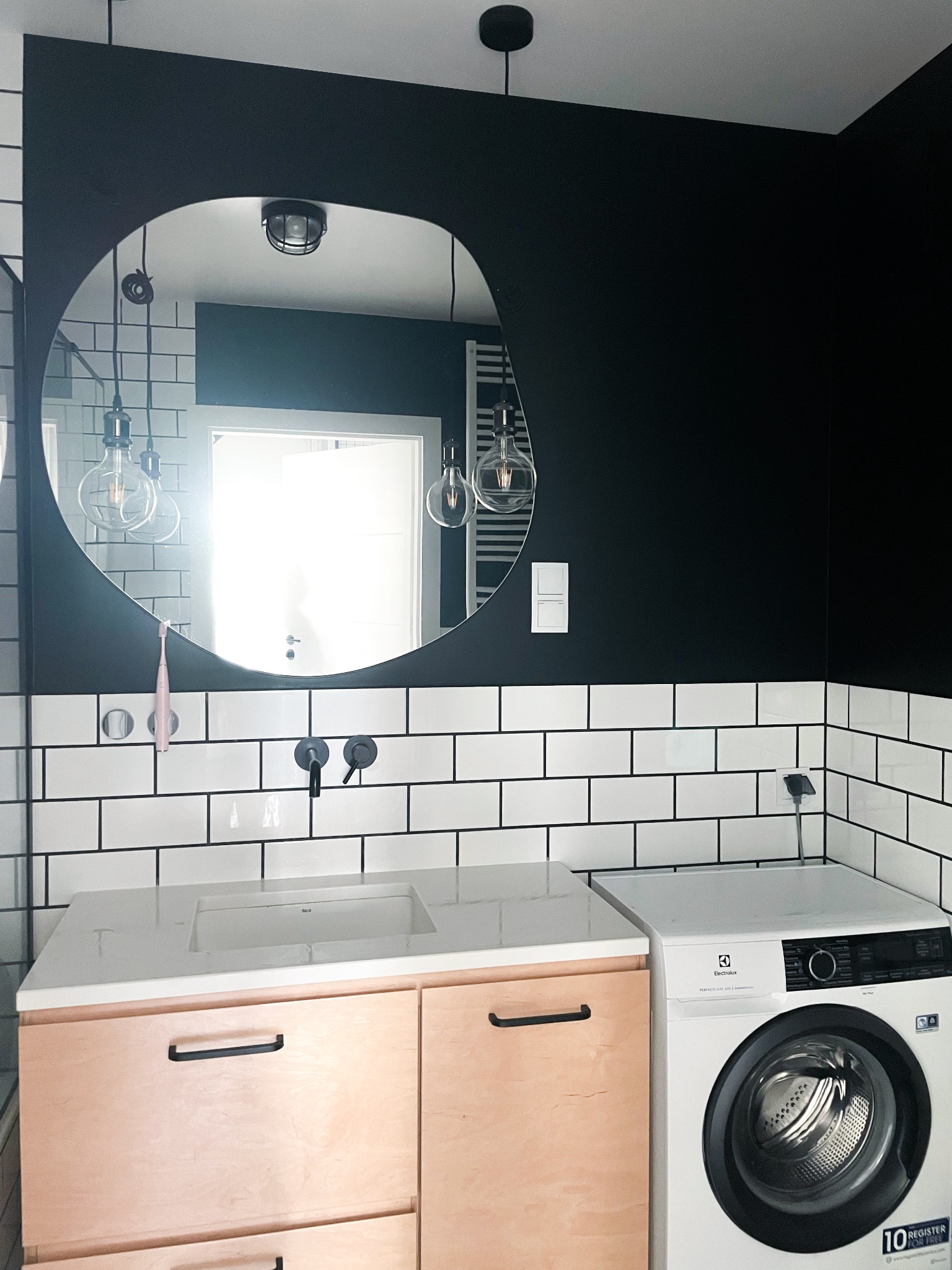 Czarno-biała łazienka z okrągłym lustrem, białymi płytkami typu metro z czarną fugą, drewnianą szafką pod umywalką, czarną ścianą i pralką Electrolux.