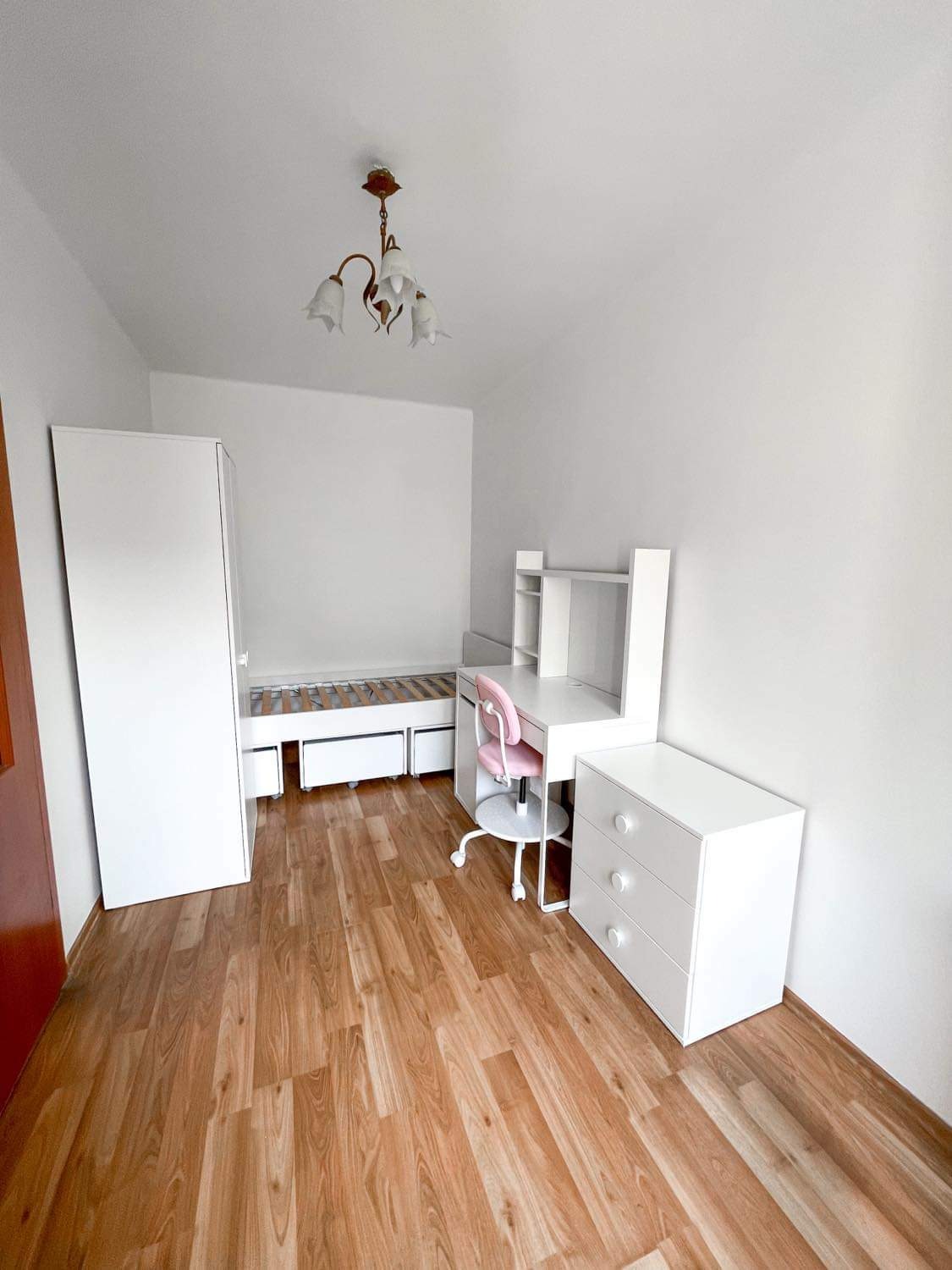Jasny pokój z drewnopodobną podłogą, białymi meblami: łóżko z szufladami, biurko z nadstawką, komoda i różowe krzesło obrotowe. Na suficie lampa z białymi kloszami.