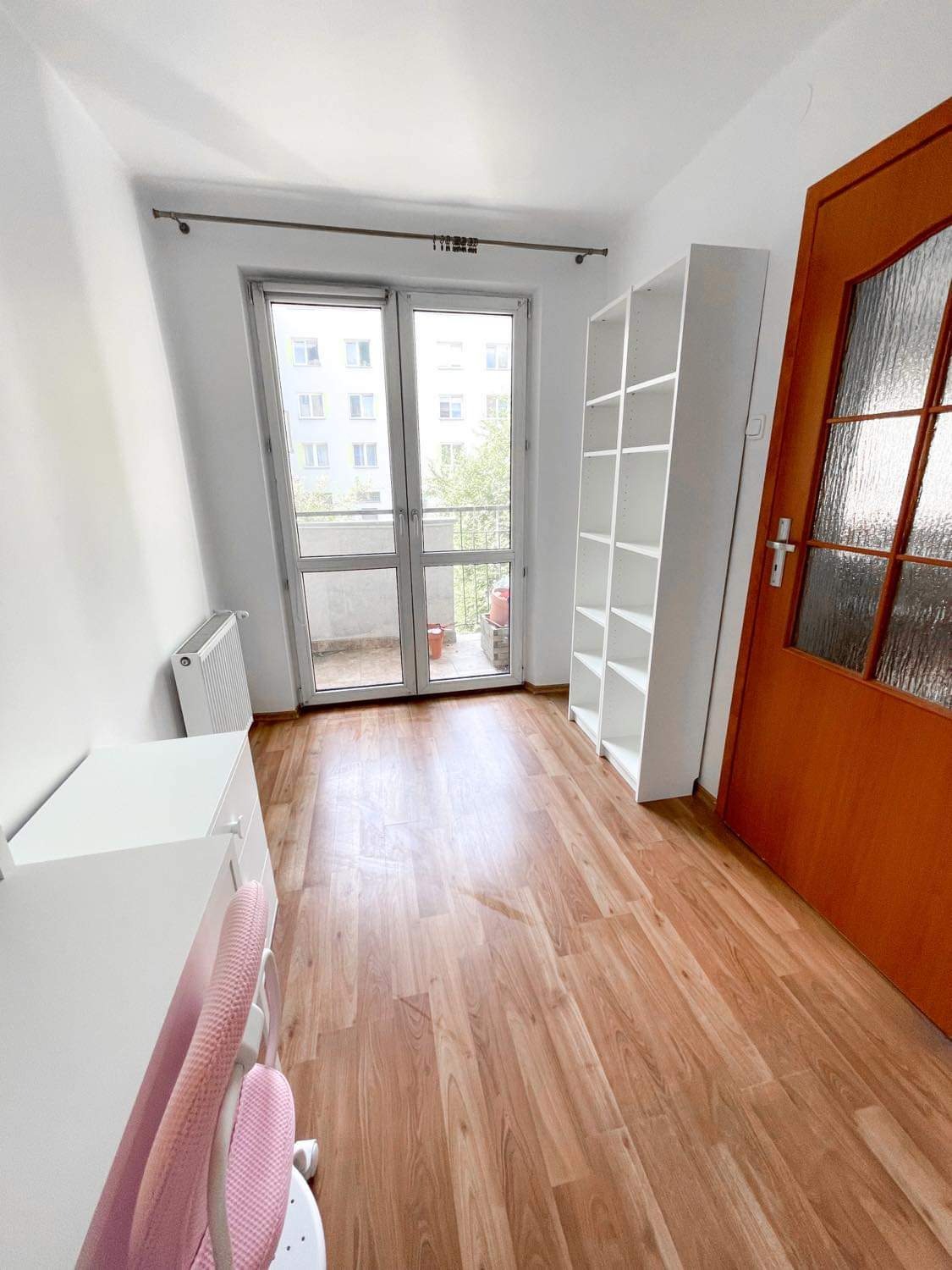 Jasne wnętrze pokoju z panelami podłogowymi, białymi ścianami, balkonem i pomarańczowymi drzwiami, widok na biały regał i grzejnik.