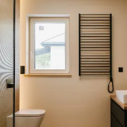Jasna łazienka z drewnianą podłogą i czarnym grzejnikiem drabinkowym. Umywalka z drewnianym blatem, toaleta pod oknem. Minimalistyczny design.