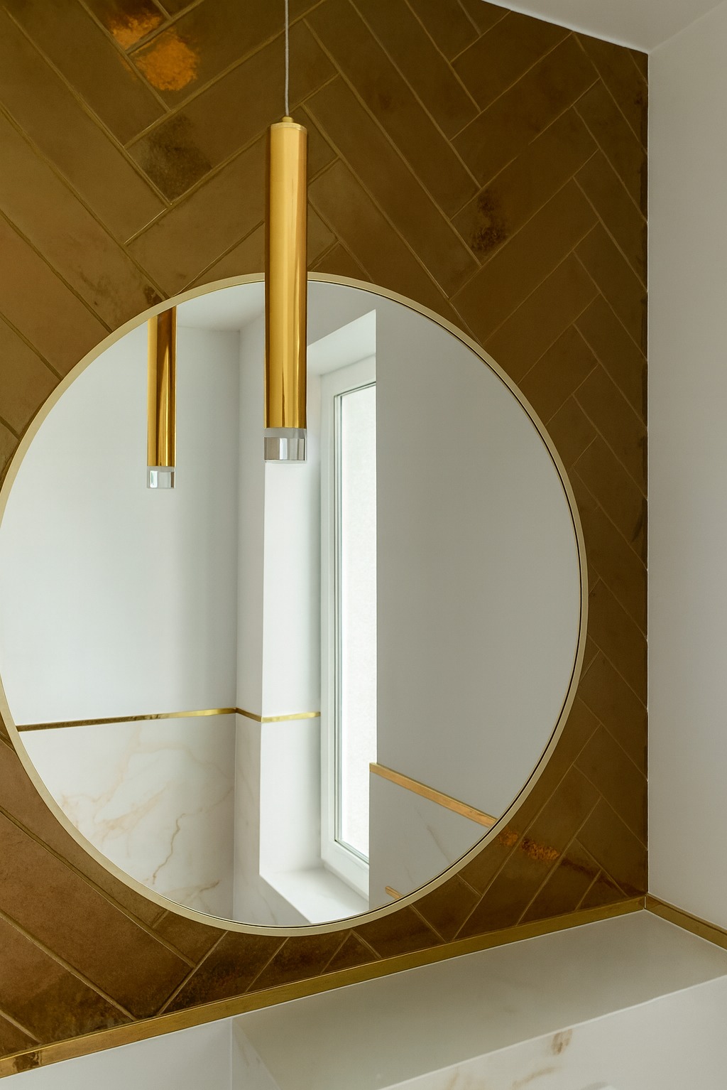 Łazienka w złotej tonacji: ściana z płytek ułożonych w jodełkę, okrągłe lustro z złotą ramą i wisząca lampa. Widoczne marmurowe elementy wykończenia.