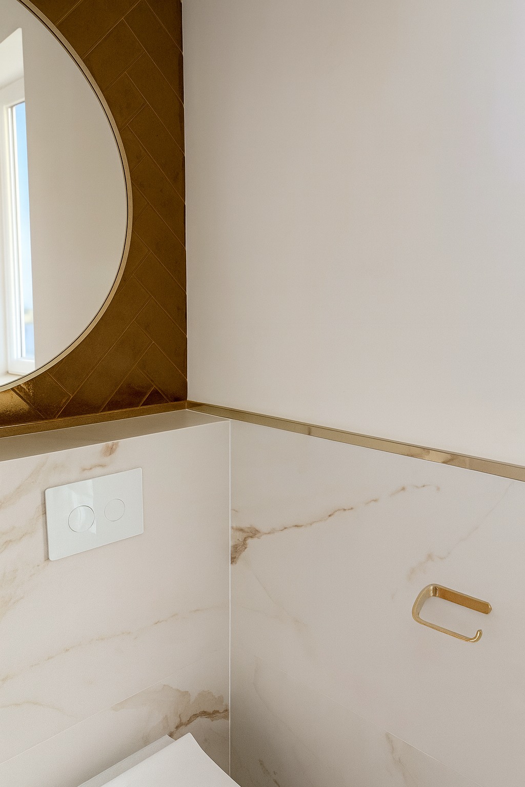 Elegancka łazienka z okrągłym lustrem i złotymi akcentami. Płytki imitujące marmur, biała armatura i minimalistyczny design tworzą spójną całość.