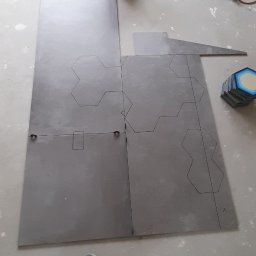 Your Best Dom IHOR SMOLIARSKYI - Przygotowane do ułożenia szare płytki podłogowe z naniesionym wzorem heksagonów, widoczne rury instalacyjne w tle, kartonowe pudełko i fragment stroju roboczego