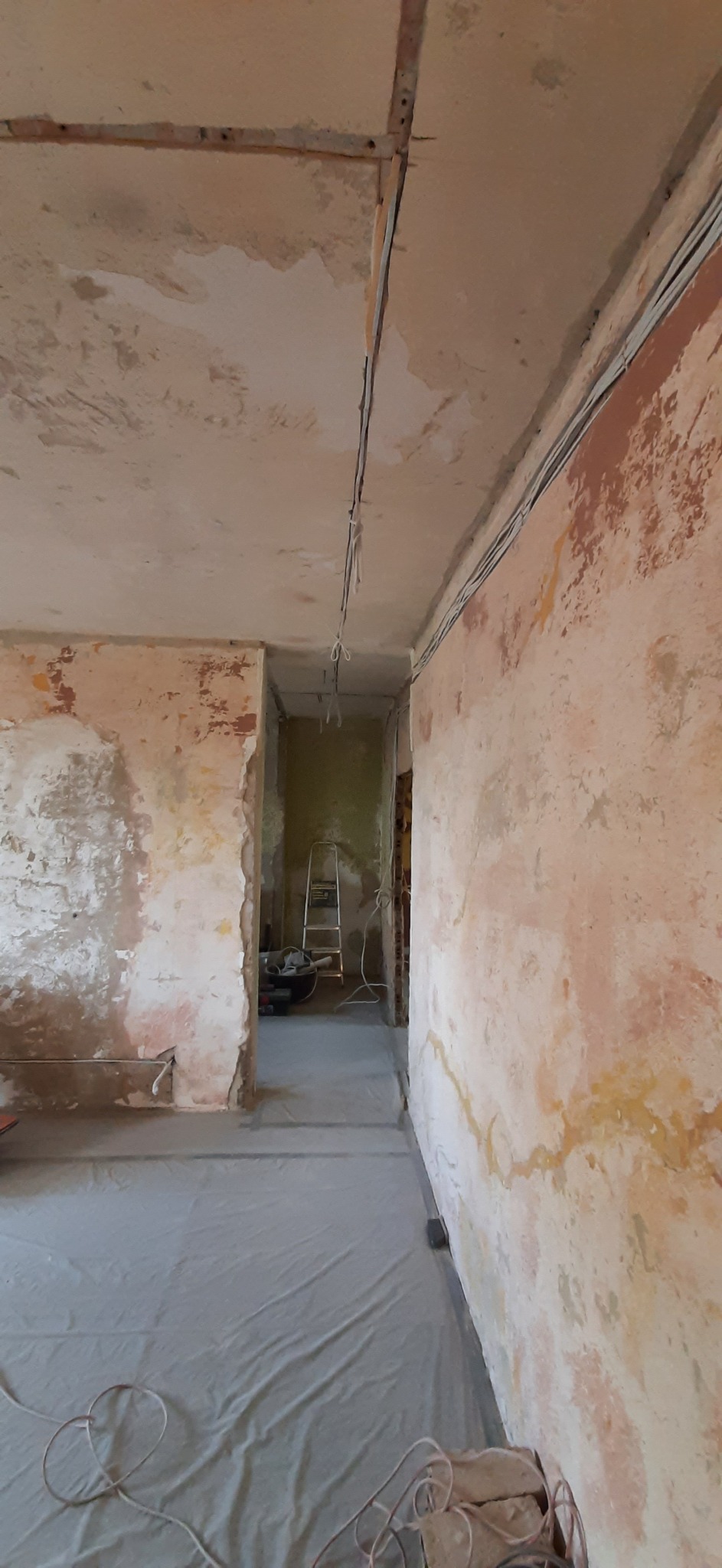 Pokój w trakcie przygotowania do prac wykończeniowych: zabezpieczona podłoga folią, ściany ze śladami starej farby i tynku, widoczne kable elektryczne, drabina w przejściu do kolejnego pomieszczenia.