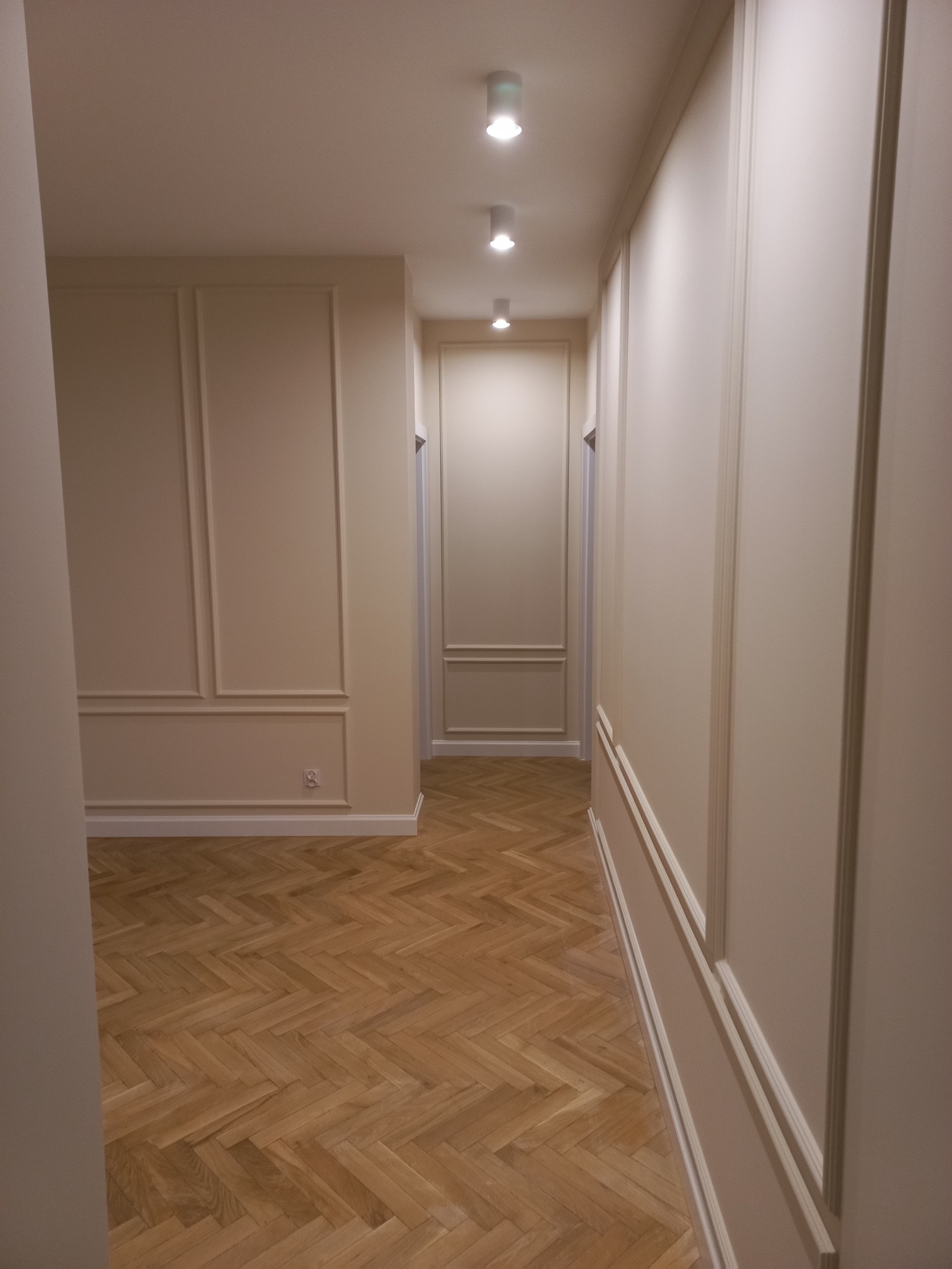 Korytarz w jasnych barwach z parkietem ułożonym w jodełkę, ozdobne panele ścienne z listew, oświetlenie punktowe w suficie.