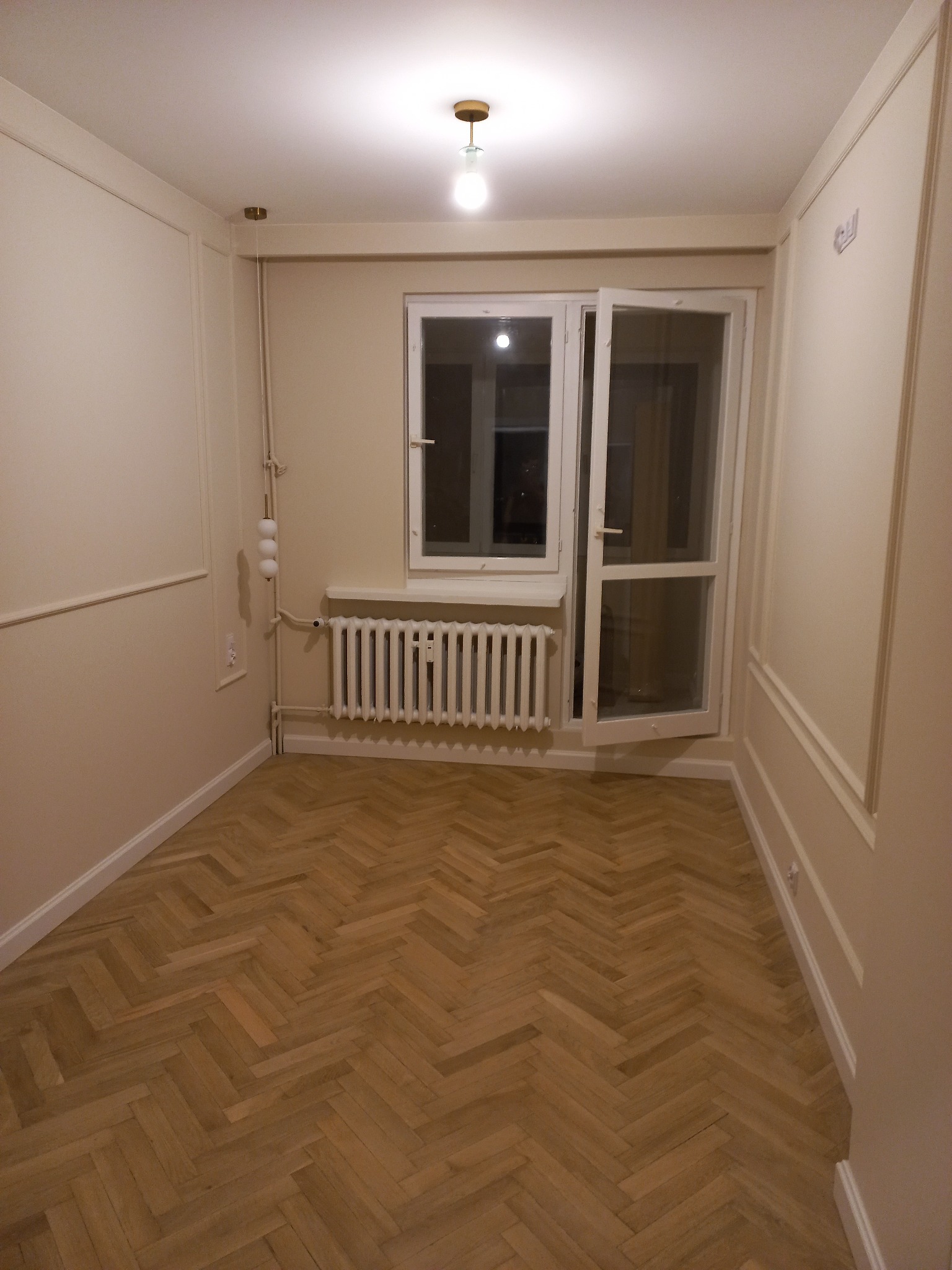 Pusty pokój z parkietem ułożonym w jodełkę, świeżo pomalowane ściany w kolorze kremowym z dekoracyjnymi listwami, otwarte białe drzwi balkonowe i grzejnik.