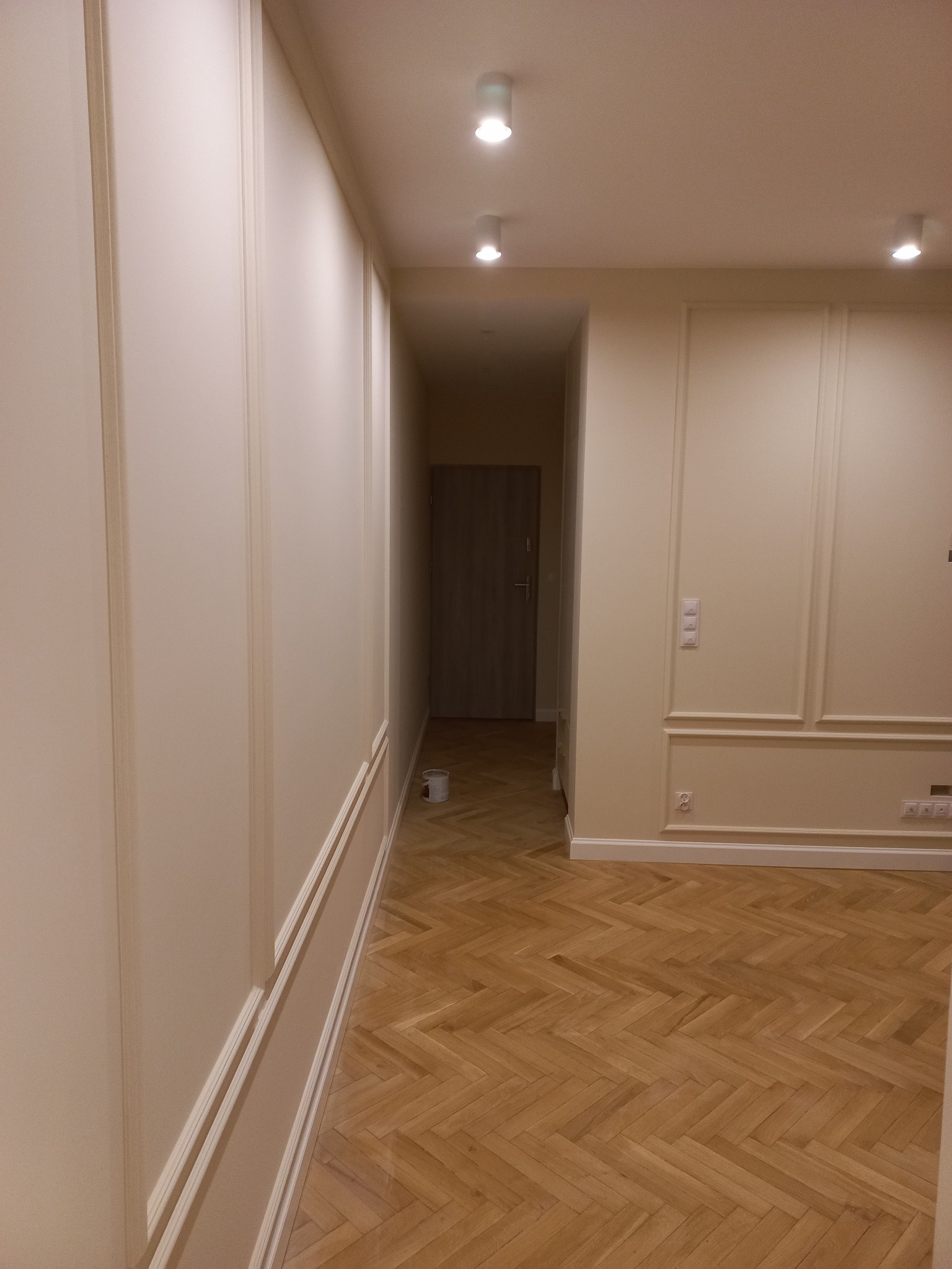Kremowe ściany z dekoracyjnymi listwami w odnowionym wnętrzu, parkiet w jodełkę, widoczny kubełek z farbą w przejściu do innego pomieszczenia.