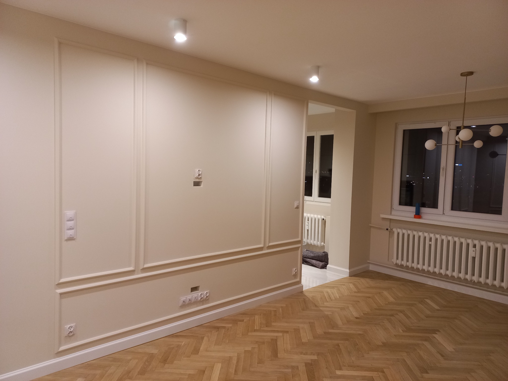 Jasne, przestronne wnętrze po remoncie z parkietem w jodełkę, kremowymi ścianami z dekoracyjnymi listwami, nowoczesnym oświetleniem i widokiem na miasto za oknem.