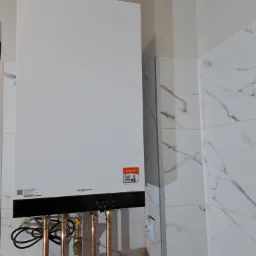 Nowoczesny, biały piec gazowy Viessmann zamontowany na ścianie wyłożonej marmurkowymi płytkami, z widocznymi miedzianymi rurami przyłączeniowymi i elementami instalacji.