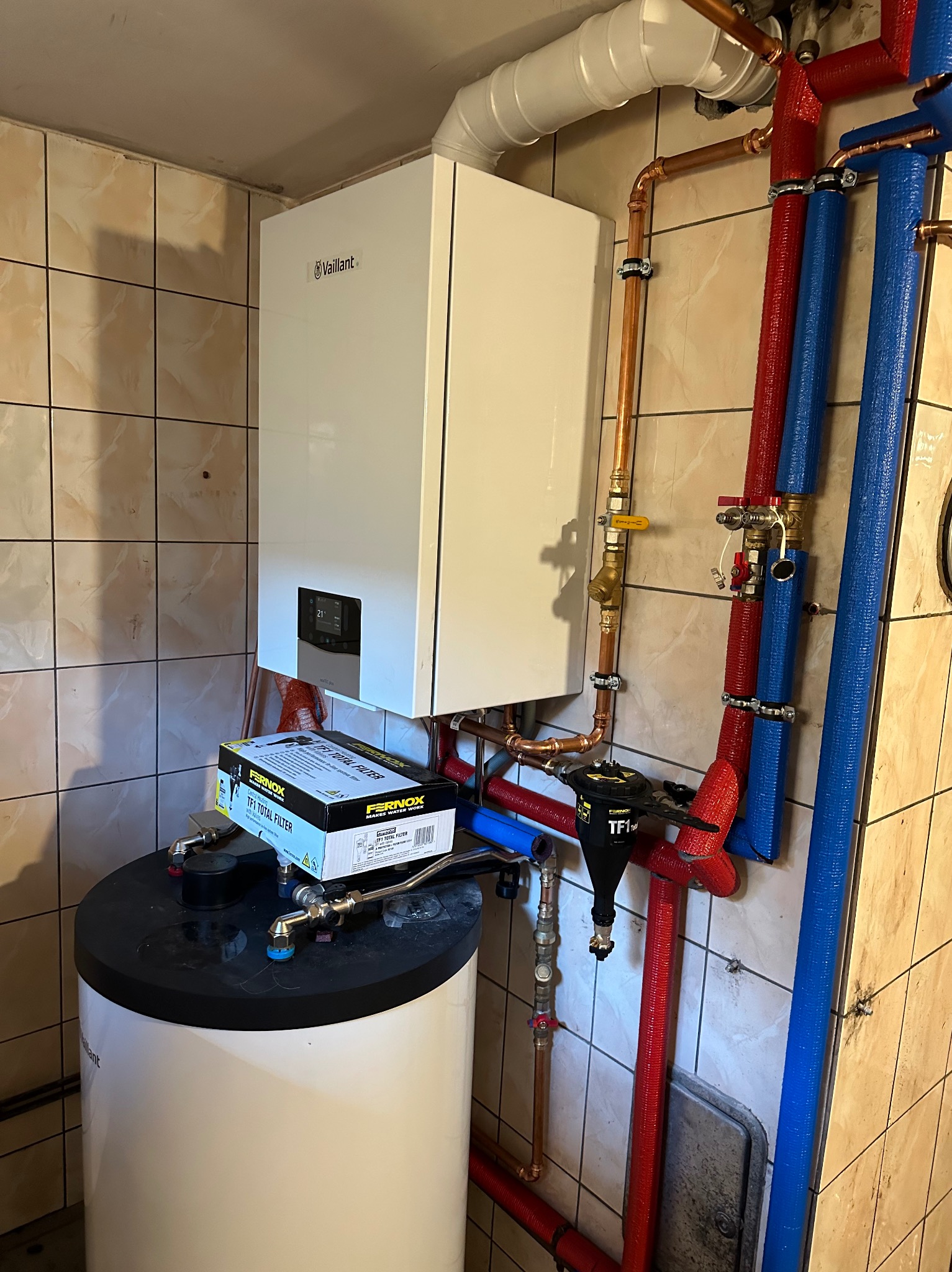 Biały wiszący kocioł gazowy Vaillant z podłączonymi rurami miedzianymi i izolowanymi rurami w kolorach czerwonym i niebieskim, zamontowany na ścianie z beżowych płytek, z filtrem magnetycznym...