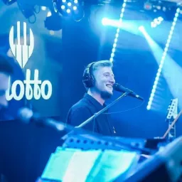 Wokalista z uśmiechem śpiewa do mikrofonu w słuchawkach na tle logotypu LOTTO i niebieskiego oświetlenia scenicznego, widoczny klawiszowiec i gitara.