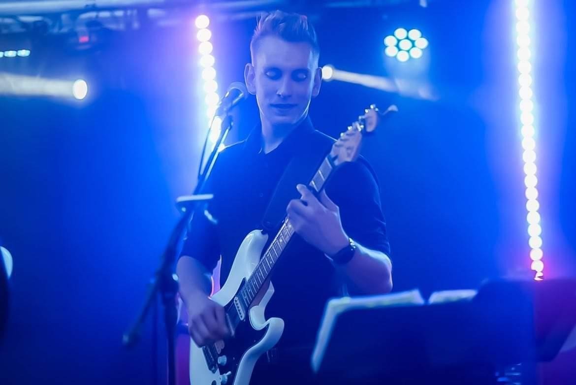 Gitarzysta w czarnej koszuli gra na białej gitarze elektrycznej na scenie oświetlonej niebieskim światłem, z mikrofonem i pulpitem na nuty w pobliżu.