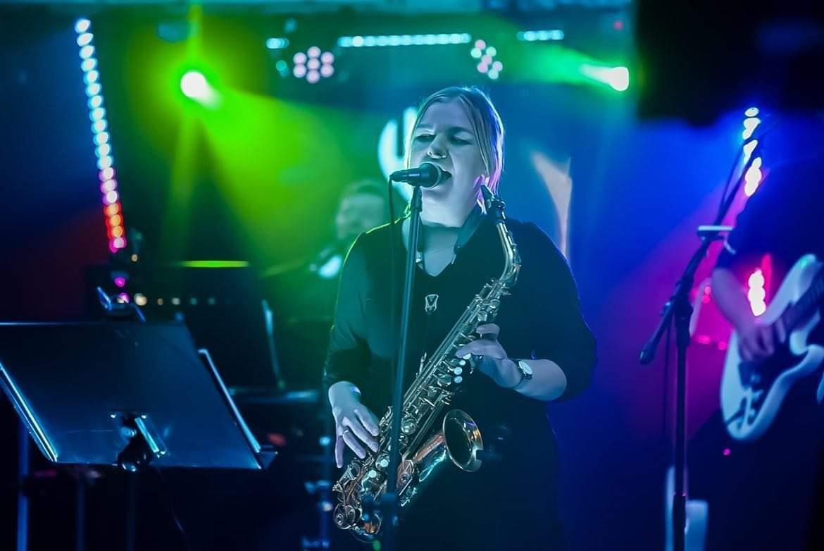 Kobieta z saksofonem śpiewa do mikrofonu na scenie, obok widoczny gitarzysta, oświetlenie sceniczne w kolorach zielonym i niebieskim.