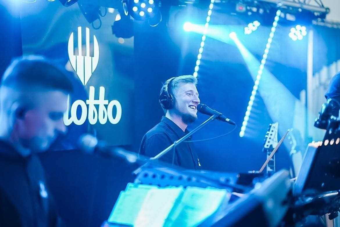 Wokalista z uśmiechem śpiewa do mikrofonu w słuchawkach na tle logotypu LOTTO i niebieskiego oświetlenia scenicznego, widoczny klawiszowiec i gitara.