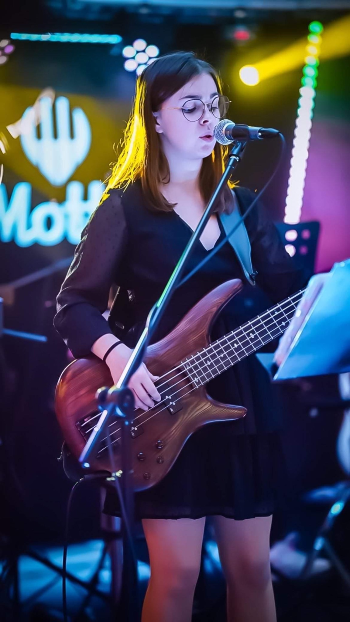Młoda kobieta w okularach śpiewa do mikrofonu i gra na basie na scenie z kolorowym oświetleniem.
