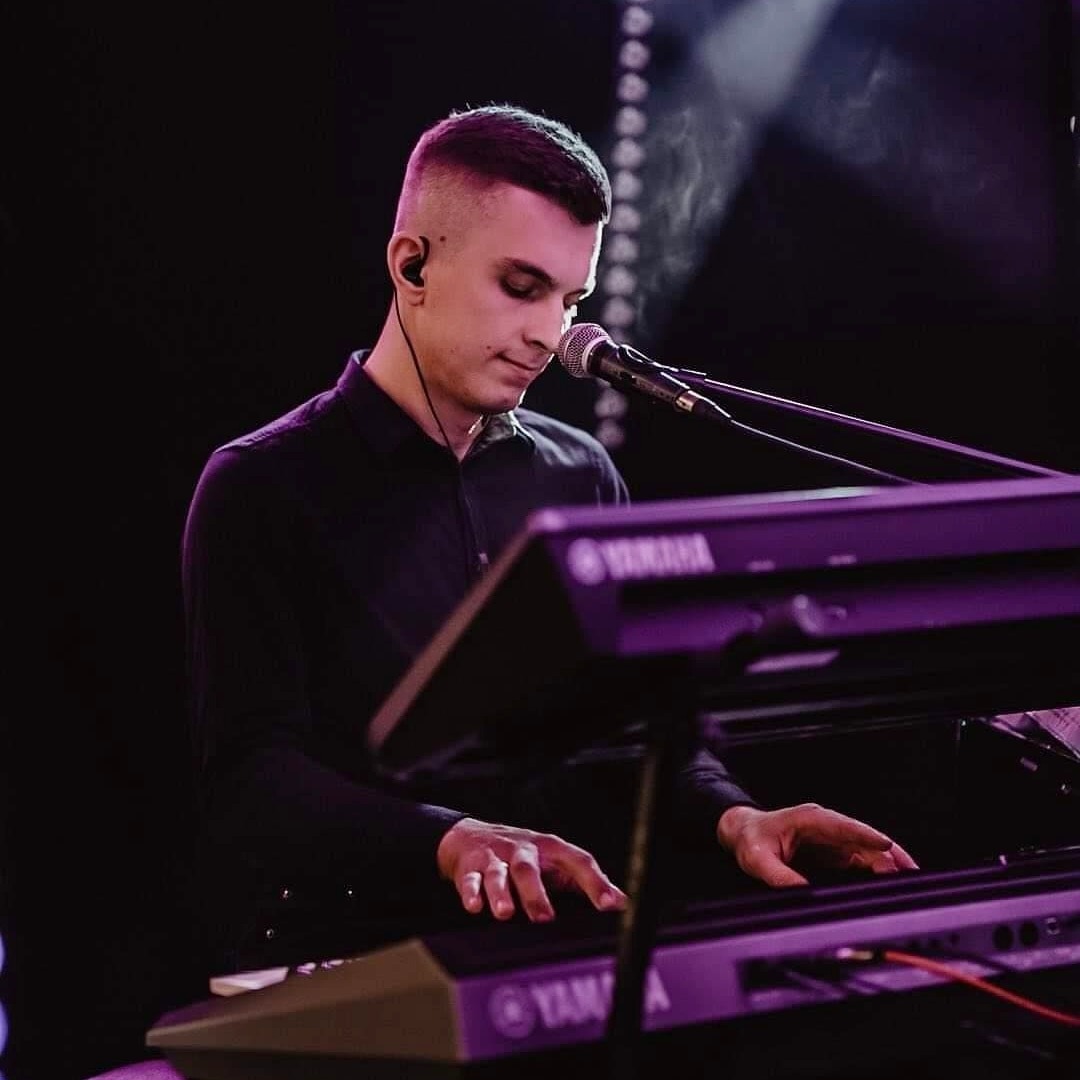 Młody muzyk w czarnej koszuli gra na keyboardzie Yamaha podświetlonym fioletowym światłem, z mikrofonem blisko ust i słuchawką w uchu, podczas występu na scenie.