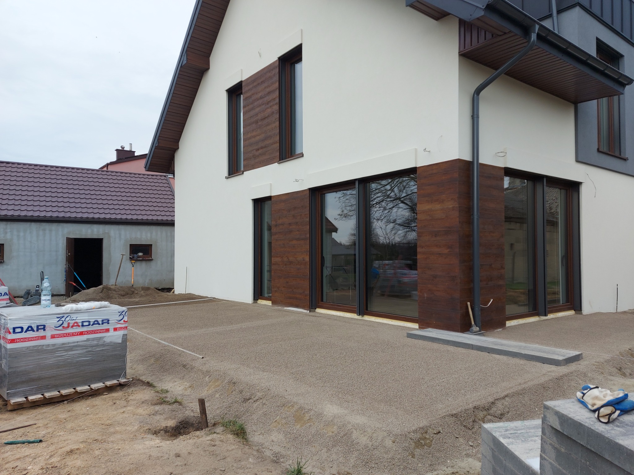 Przygotowany podkład z kruszywa pod taras z kostki brukowej przed nowoczesnym domem z drewnianą okładziną, w tle paleta z kostką brukową i budynek gospodarczy.