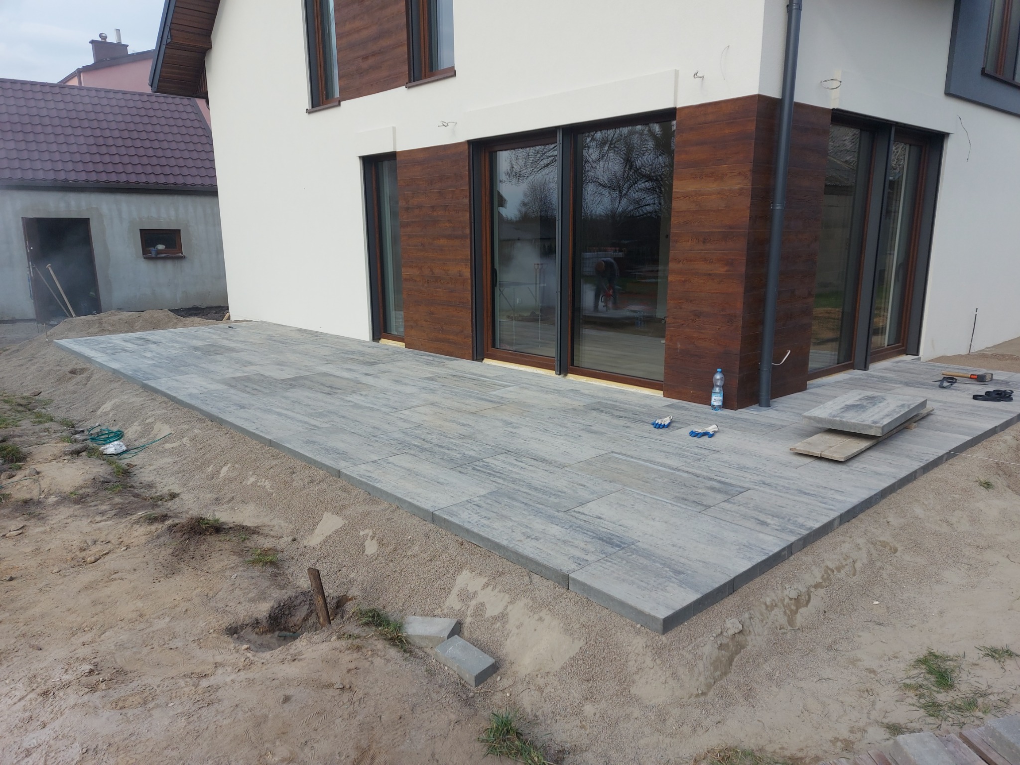 Nowo ułożony taras z szarej kostki brukowej o podłużnym kształcie, przylegający do ściany domu z drewnianymi elementami, widoczne rękawice robocze, poziomica i butelka wody mineralnej.