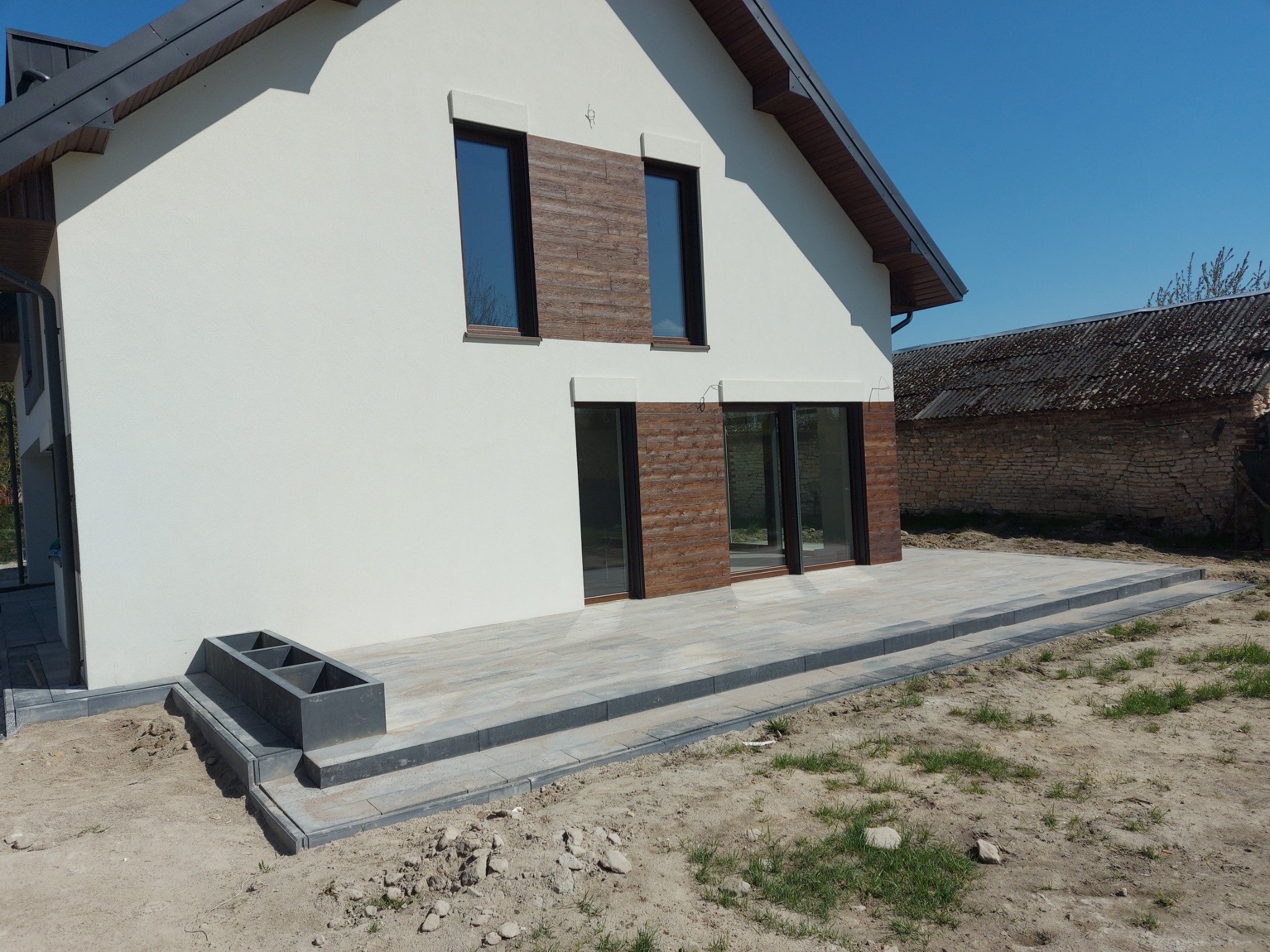 Nowoczesny dom z jasną elewacją i tarasem wyłożonym szarą kostką brukową, widoczne donice z betonu architektonicznego, w tle stary budynek gospodarczy.