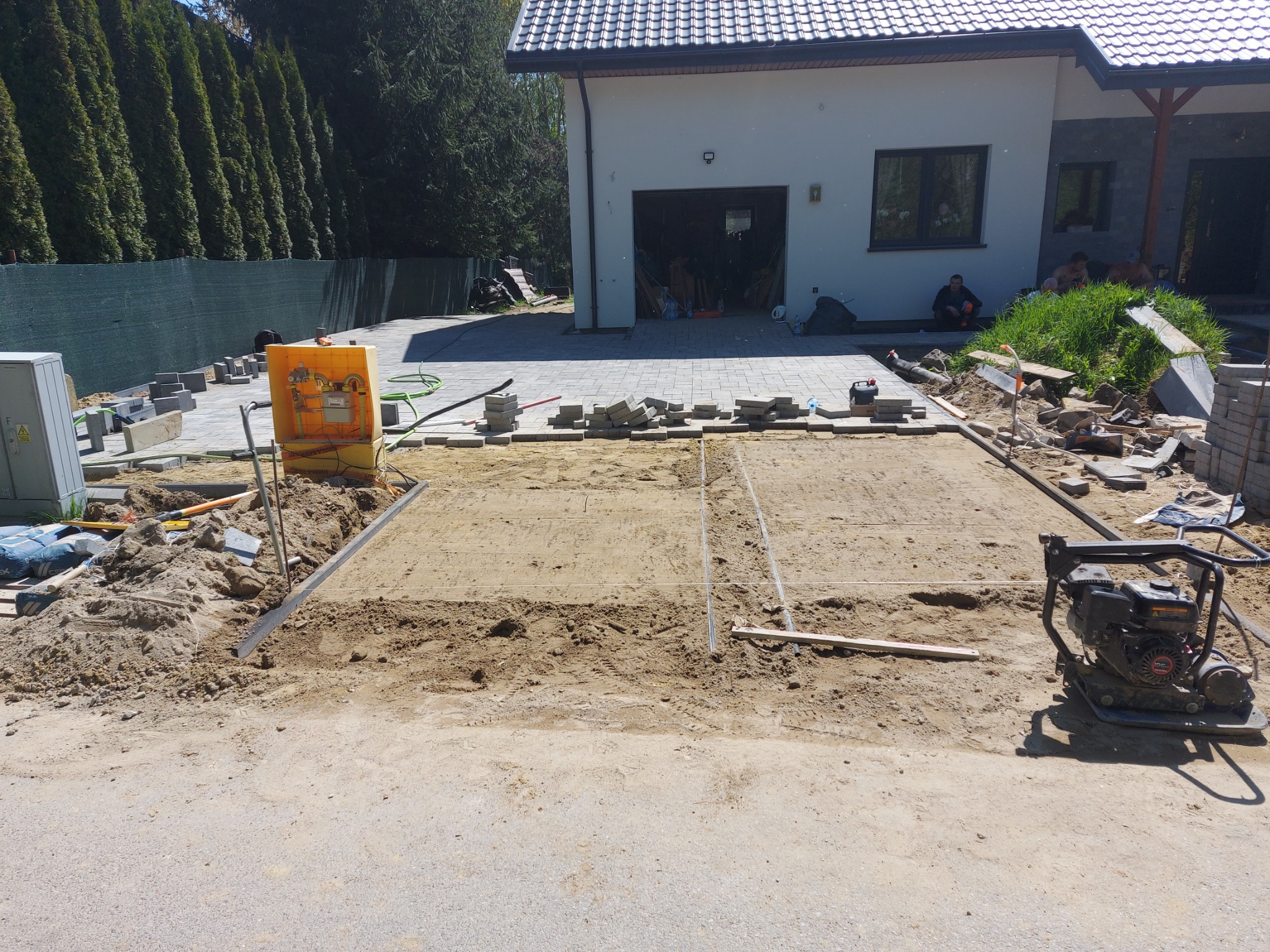 Przygotowanie podłoża pod kostkę brukową na podjeździe przed domem jednorodzinnym, widoczne narzędzia, agregat prądotwórczy, oraz stosy kostki brukowej i granitowej