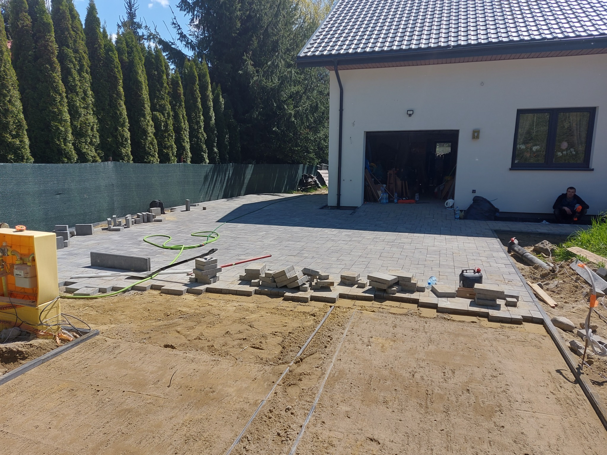 Układanie szarej kostki brukowej na podjeździe przed domem, widoczne narzędzia, piasek, częściowo ułożona nawierzchnia oraz mężczyzna siedzący w cieniu budynku.