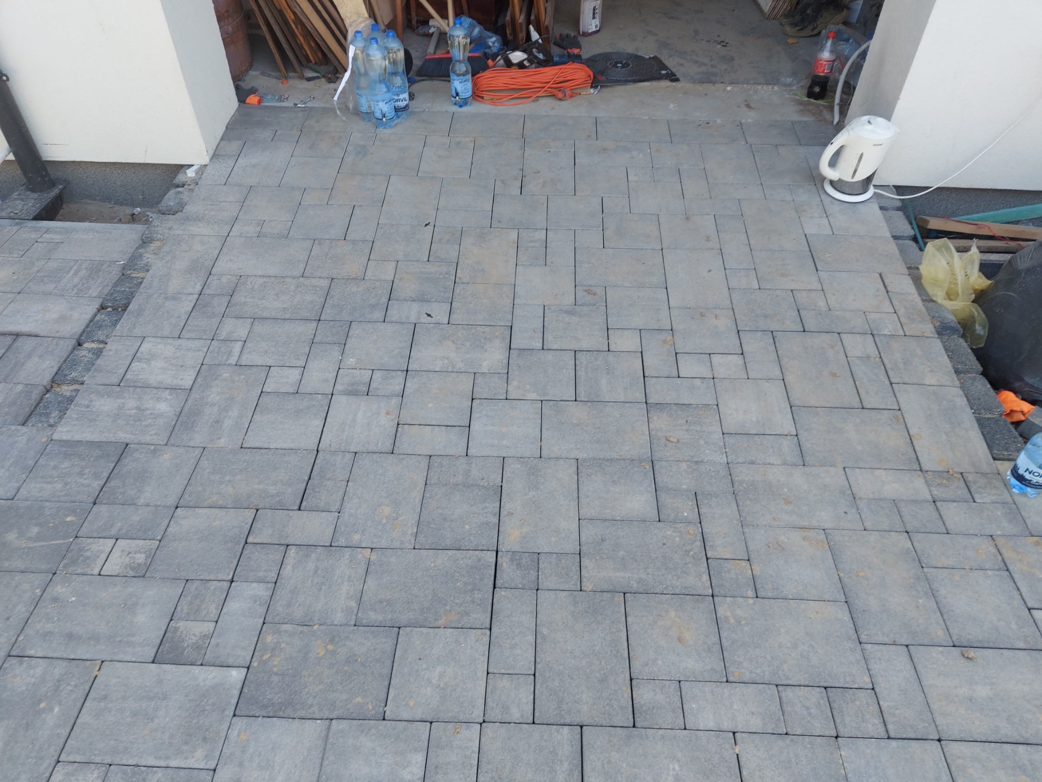 Ułożona szara kostka brukowa o różnych rozmiarach, widoczne butelki z wodą, czajnik elektryczny i rozłożony przedłużacz na tle białej ściany budynku.