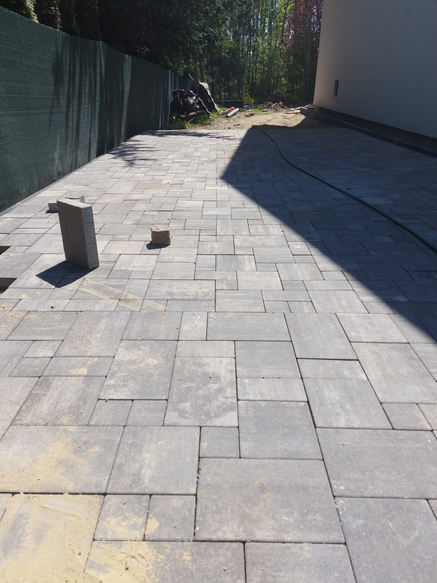 Ułożona szara kostka brukowa na podwórku, widoczne niedokończone fragmenty z piaskiem, w tle zielona siatka ogrodzeniowa i budynek w cieniu.