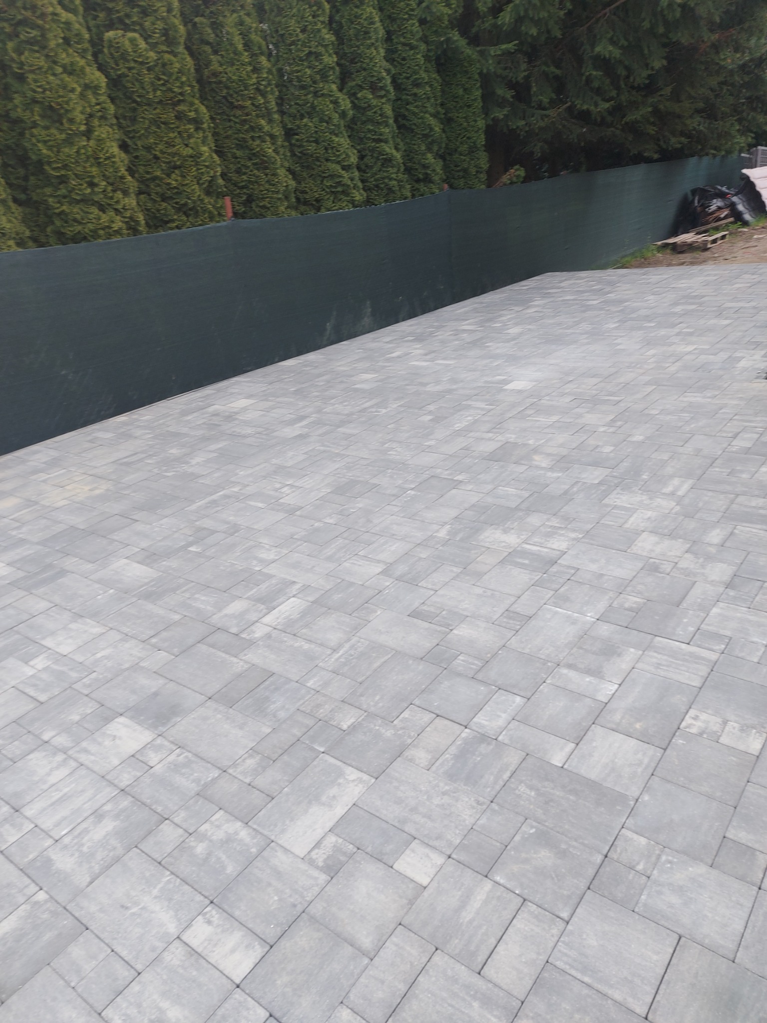 Świeżo ułożona, szara kostka brukowa tworząca równą powierzchnię, z ciemnozielonym żywopłotem w tle i materiałami budowlanymi w prawym górnym rogu.