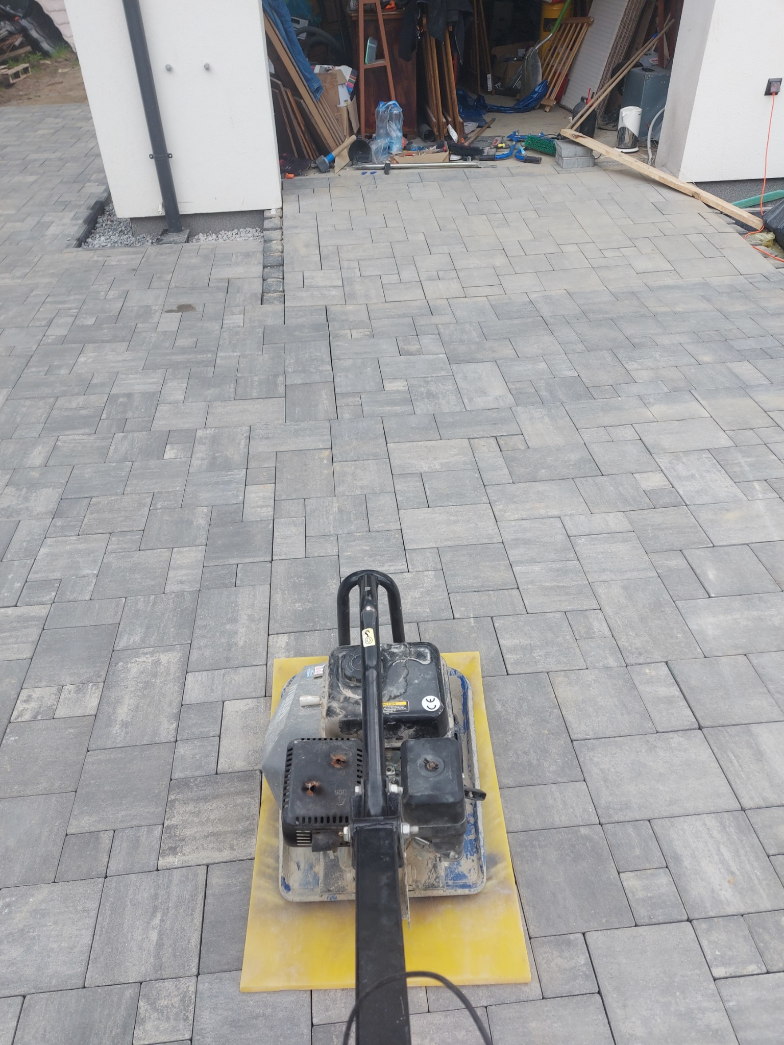 Wibro ubijak na szarej kostce brukowej ułożonej przed garażem z otwartymi drzwiami, widoczne narzędzia i materiały budowlane w tle.