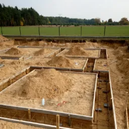 Wykop pod fundamenty domu z widocznym szalunkiem, zbrojeniem i usypanymi pryzmami piasku wewnątrz, na tle zielonego pola i lasu.