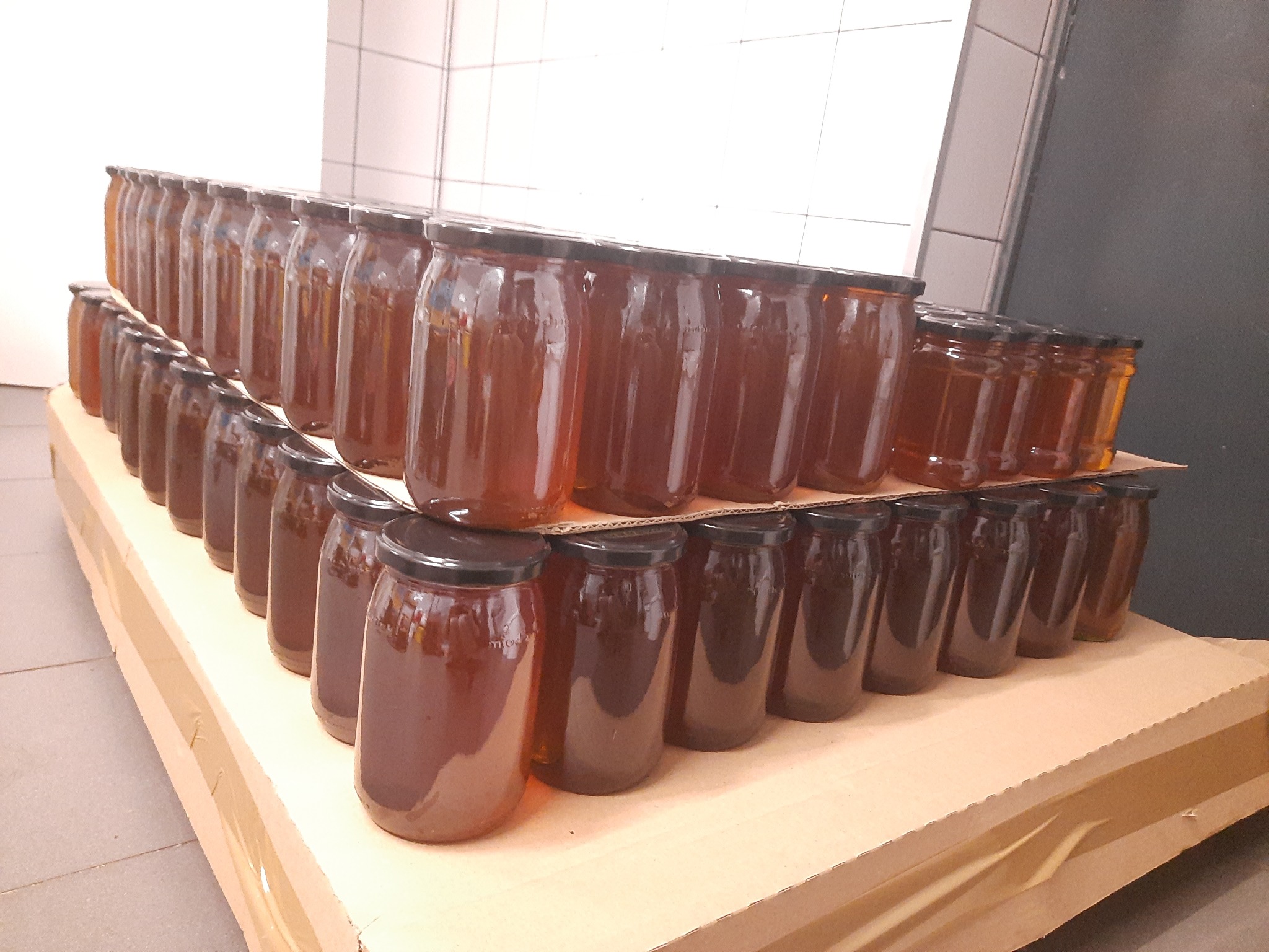 Słoiki z ciemnym płynnym produktem ułożone na kartonowym podeście w magazynie z białymi płytkami na ścianie.