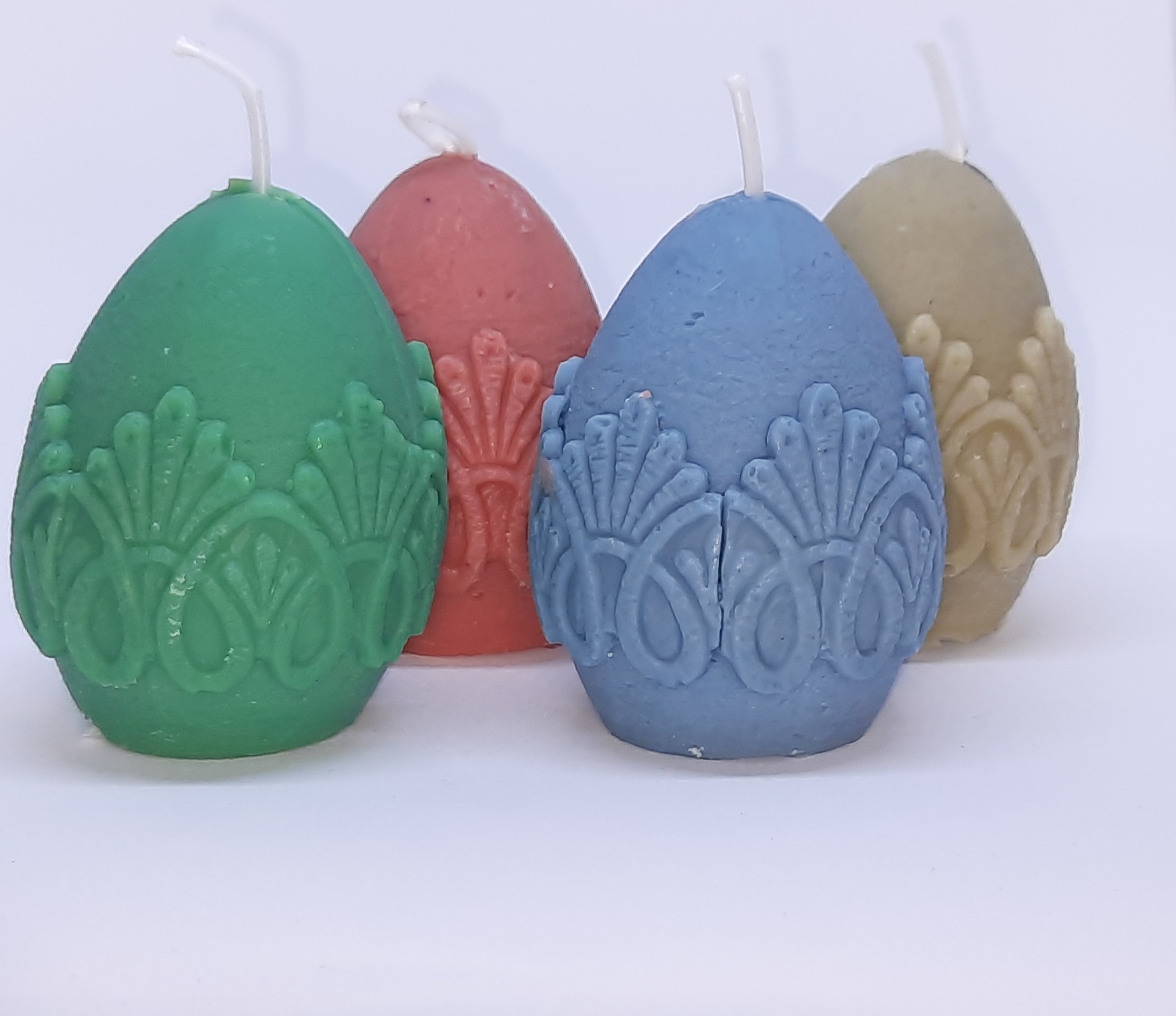 Cztery dekoracyjne świeczki w kształcie jajek, każda w innym pastelowym kolorze (zielony, różowy, niebieski, beżowy) z wytłaczanymi ornamentami, ustawione na białym tle.