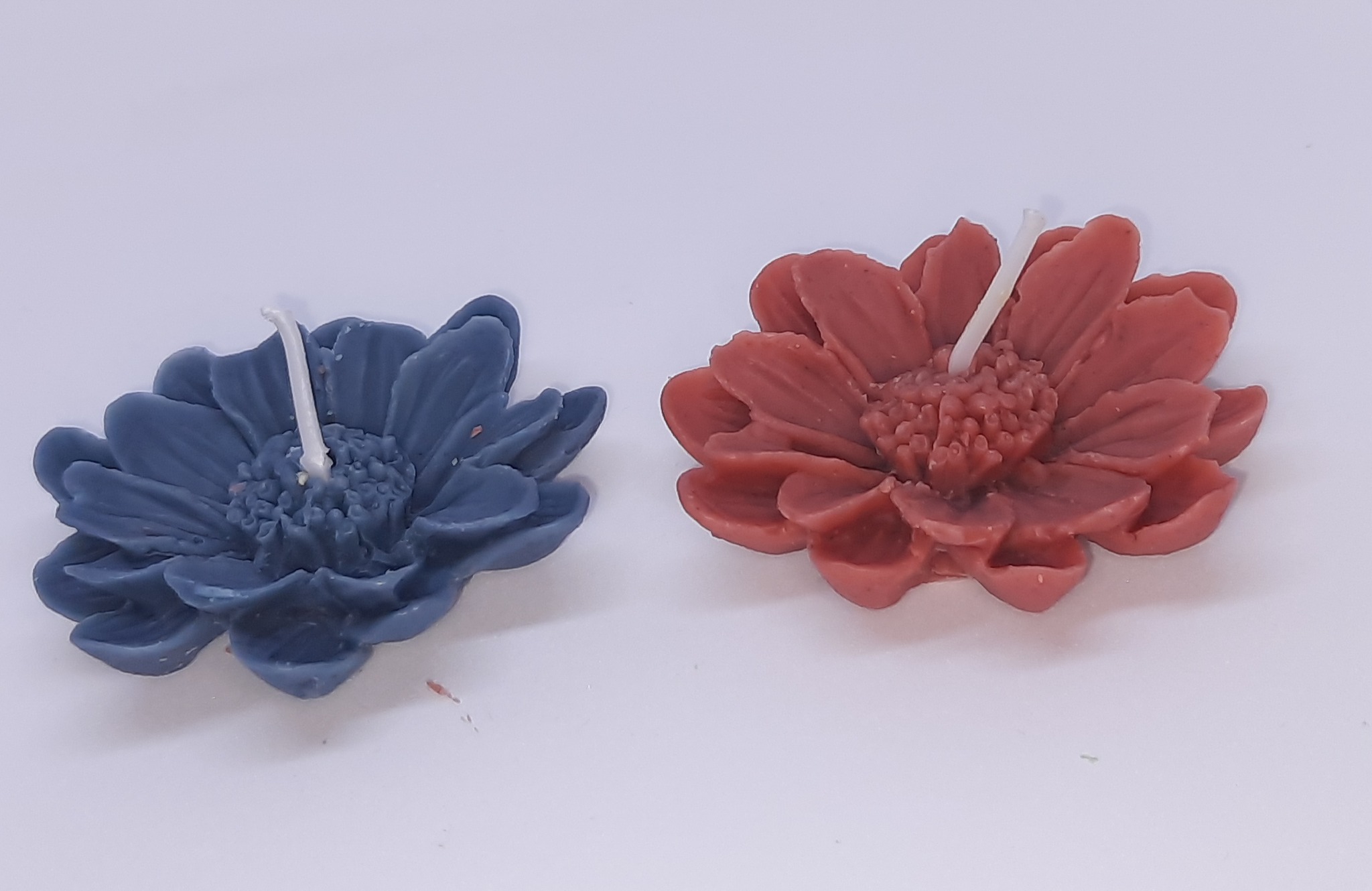 Dwie świece w kształcie kwiatów: jedna w odcieniu granatu, druga w kolorze ceglastoczerwonym, na białym tle.