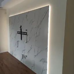 Ściana wyłożona płytkami imitującymi marmur z zamontowanym uchwytem do telewizora i podświetleniem LED po bokach, widoczne gniazdka elektryczne i fragment drewnianej podłogi.