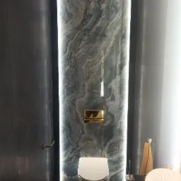 Nowoczesna łazienka z podświetlaną ścianą z szarego marmuru, wisząca toaleta z chromowanym przyciskiem, umywalka nablatowa z elementami w kolorze złota i ciemne płytki na ścianach.